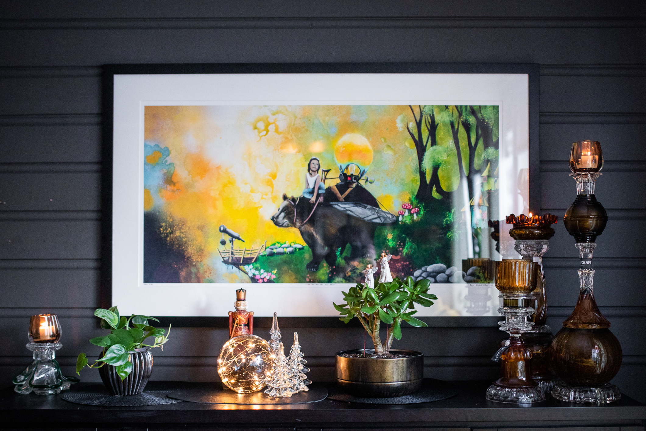 Et stort fargerikt bildet henger på veggen. På hyllen under står flere gjenbruks lysestaker. Her ser vi Ainas kunst samlet.