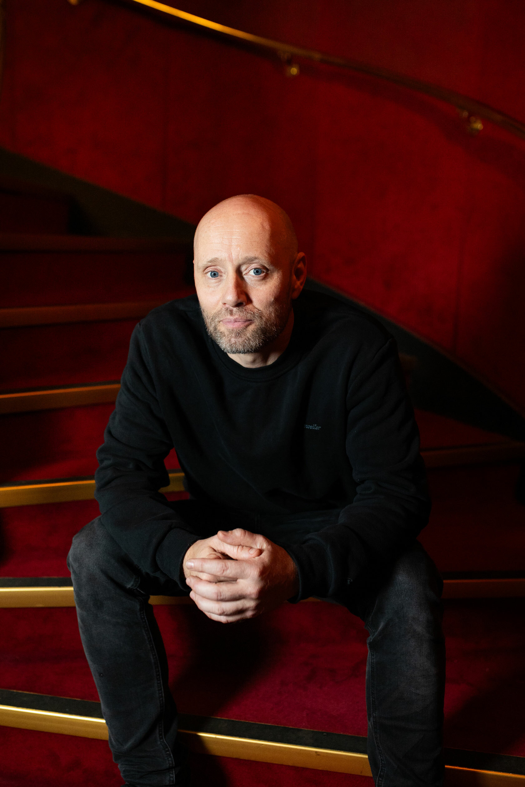 Foto: Skallet mann sitter i spiraltrapp med røde vegger og rødt teppe. Han folder hendene.