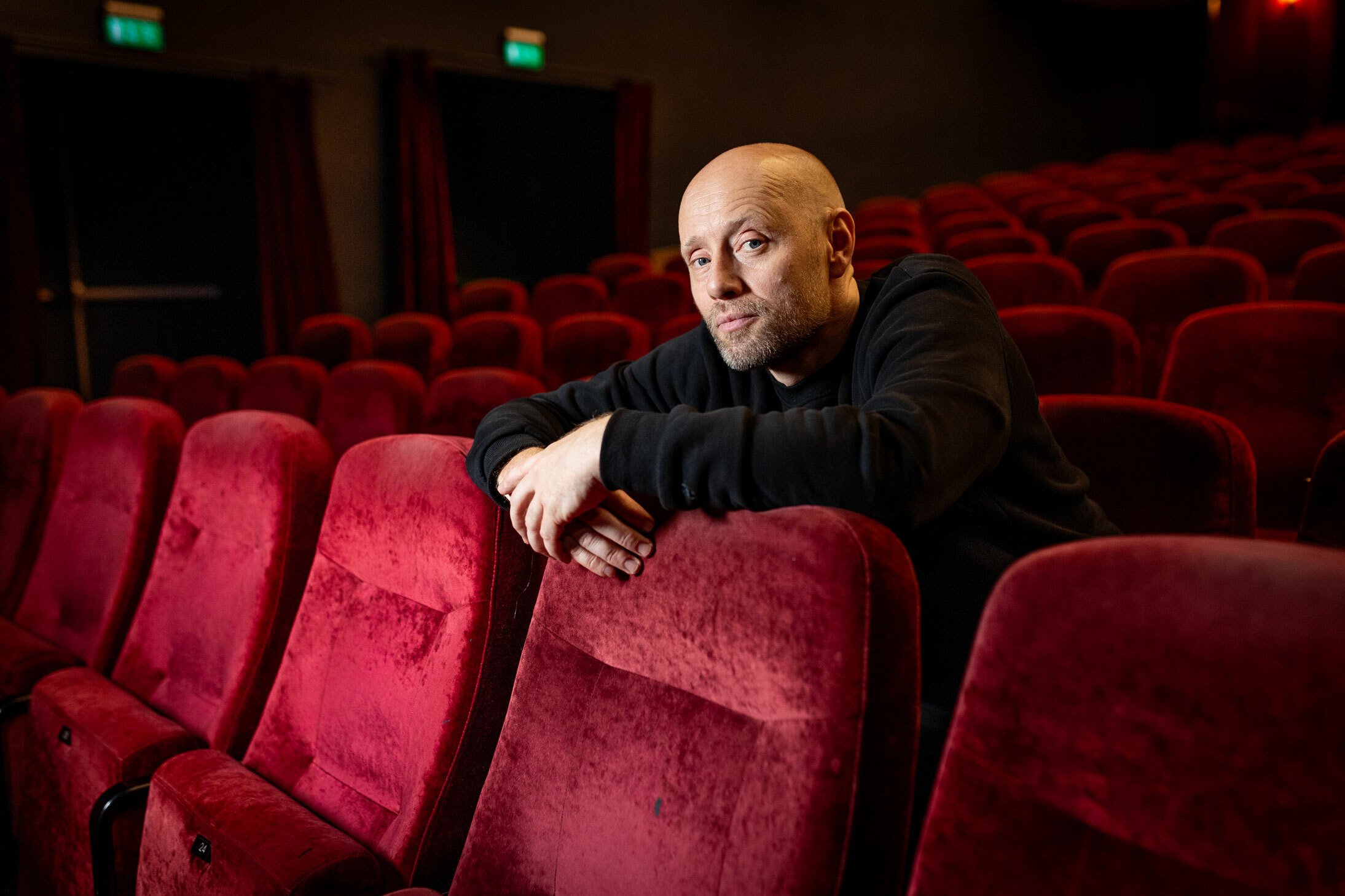 Foto: Mann uten hår sitter alene i en stor teatersal med røde plysjseter.
