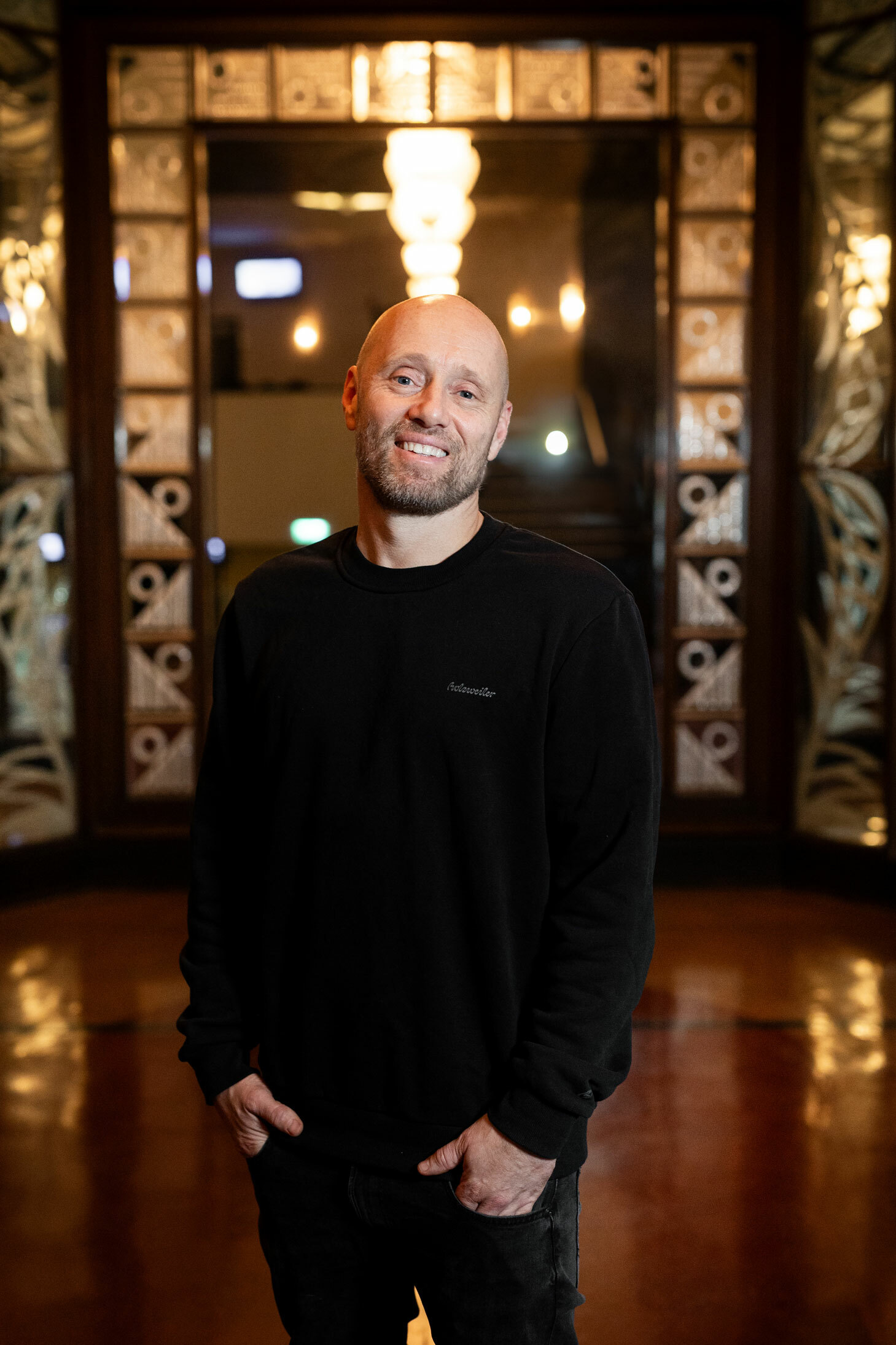 Foto: Mann uten hår smiler i en korridor med en vakker glassvegg rundt en døråpning i enden.