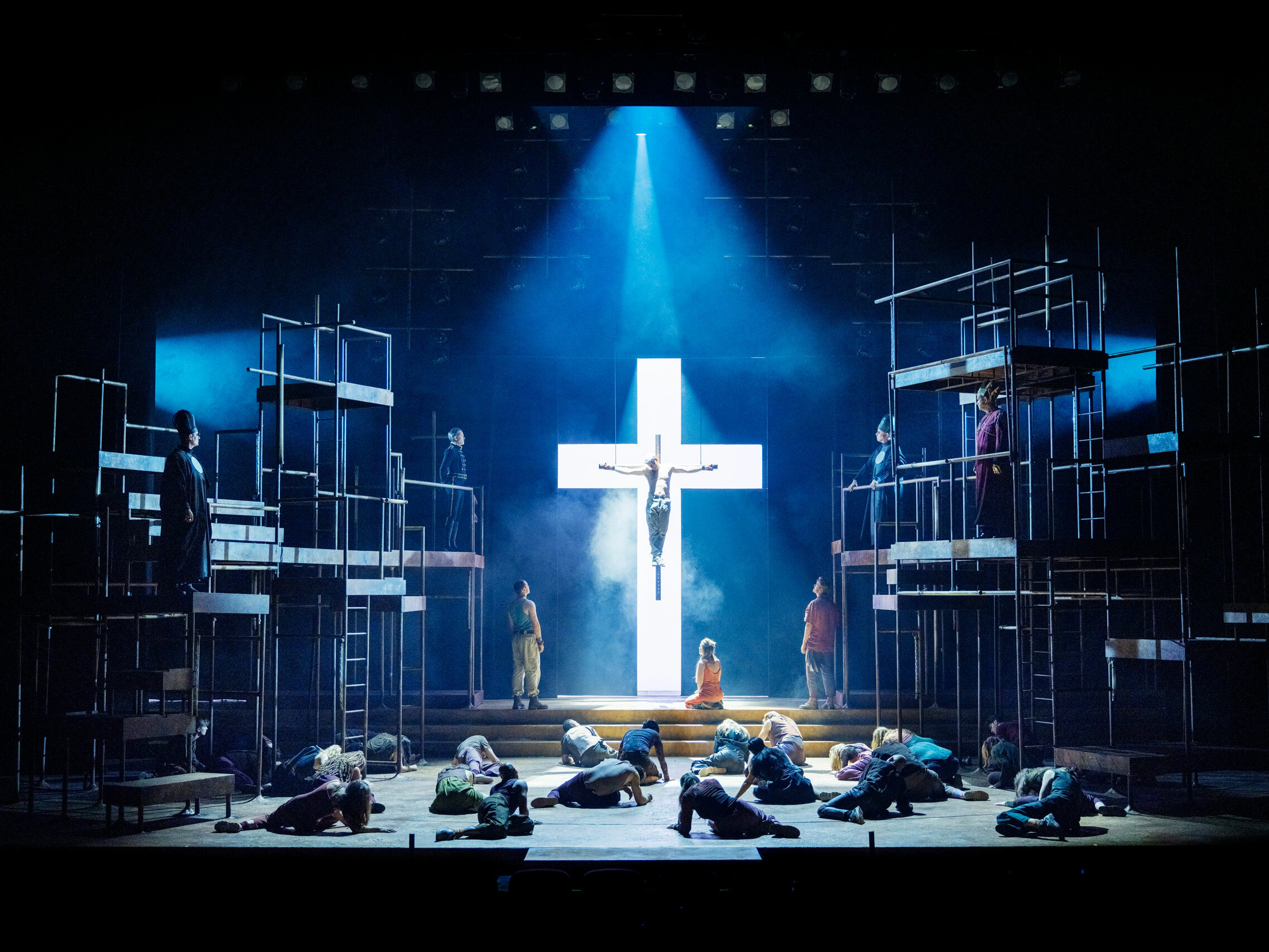 Foto: Lysende, hvitt kors med blått lys. Det henger en mann på. Foran på scenen ligger folk rundt. Det er stilaser på hver side.