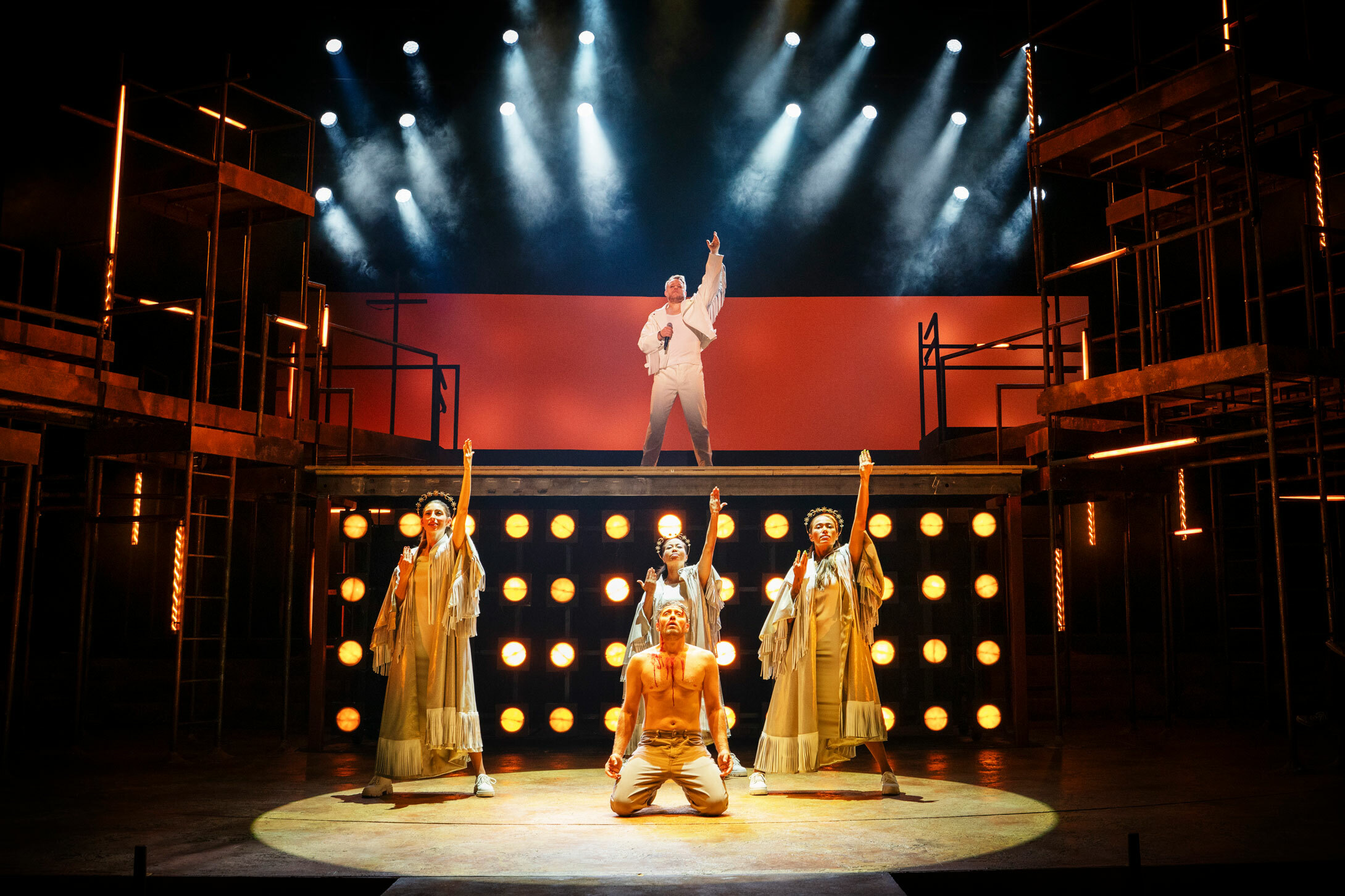 Foto: To nivåre på en scene. Nede ser vi en blødende mann med tornekrone på knærne – Jesus. På et nivå over ham ser vi en mann med hånden opp i været. Jesus-skikkelsen er omringet av tre kvinner i sølvkjoler.