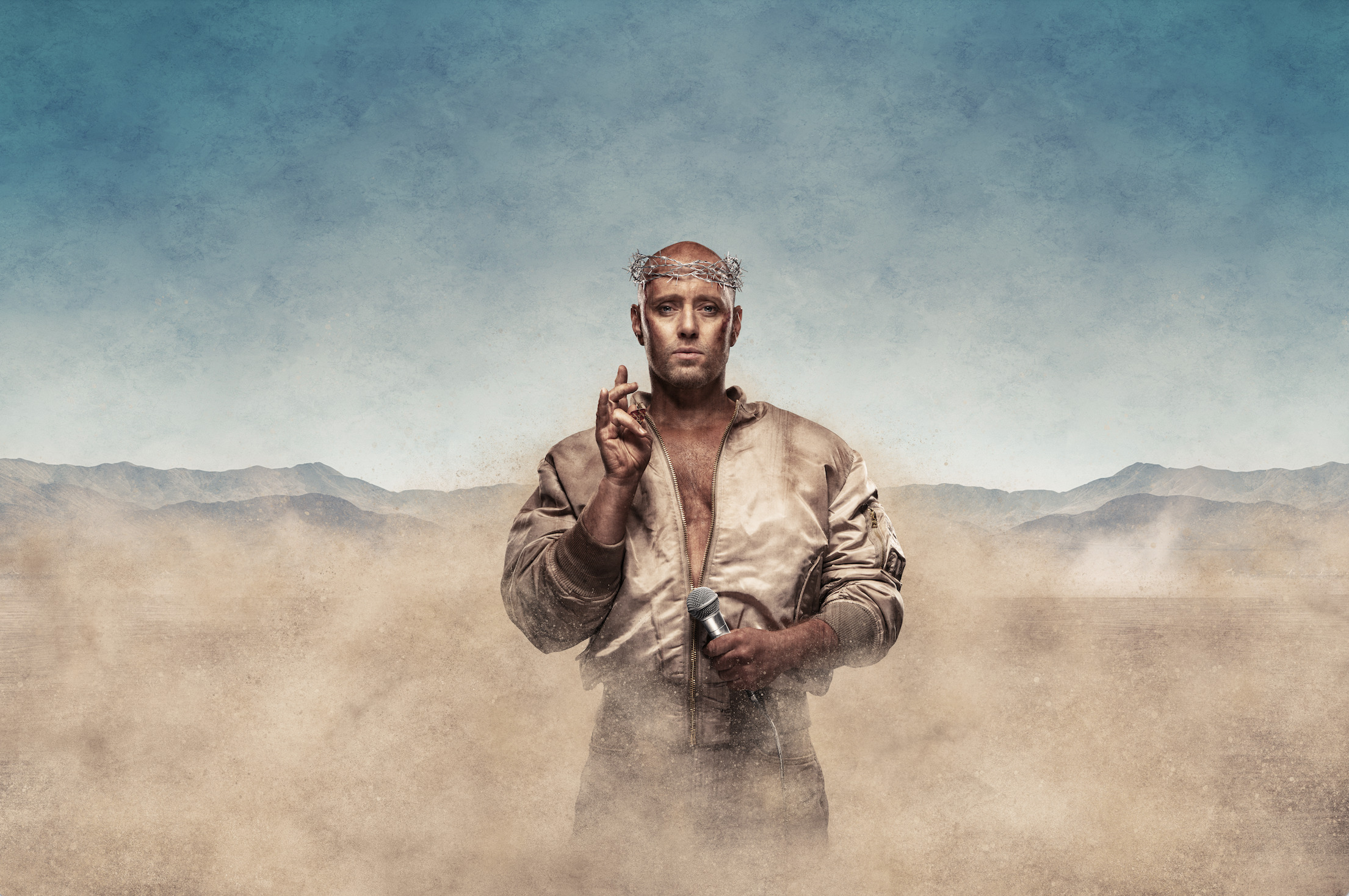 Foto: Mann i gullklær og en trornekrans på sitt skallede hode. Han står i et tørt landskap med sand og fjell-