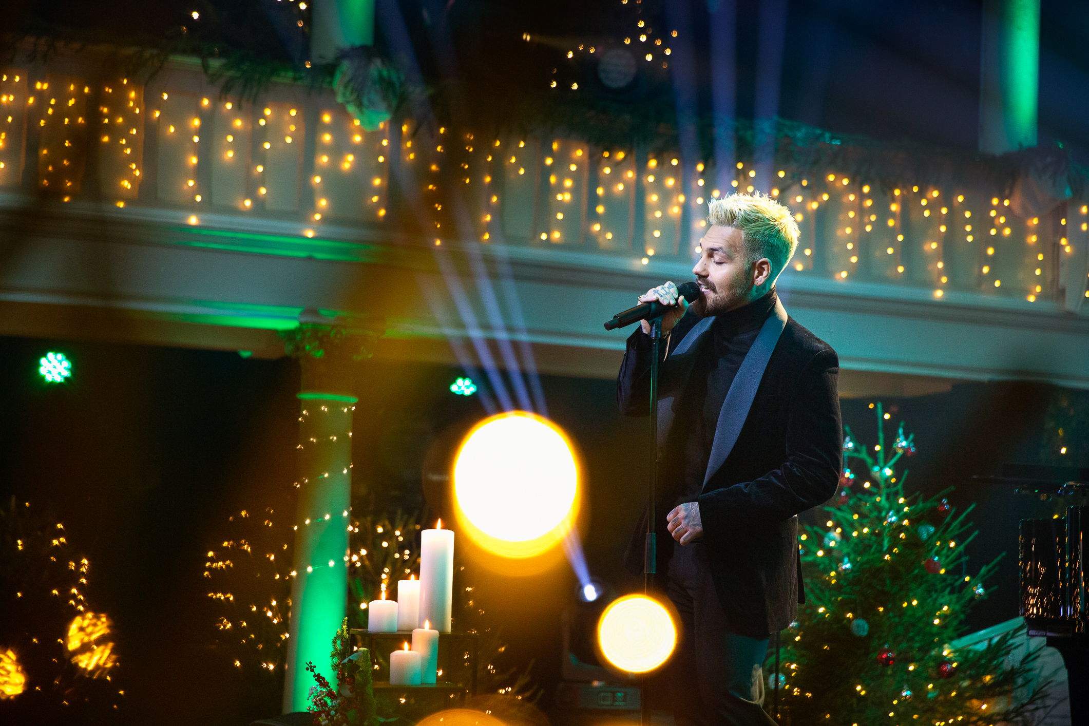 Foto: Mann med bleket hår som står og synger på en intimscene med mange gule julelys på alle kanter.