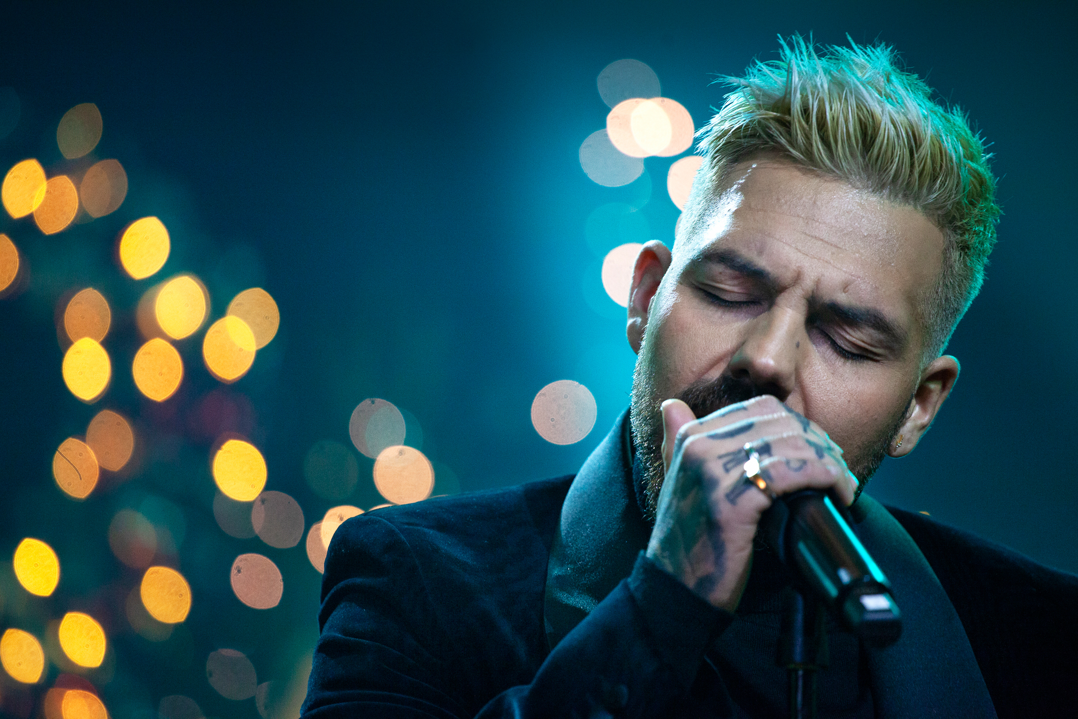 Foto: Mann med lukkede øyne som holder en tatovert hånd rundt mikrofonen mens han synger. I bakgrunnen gule scenelys.