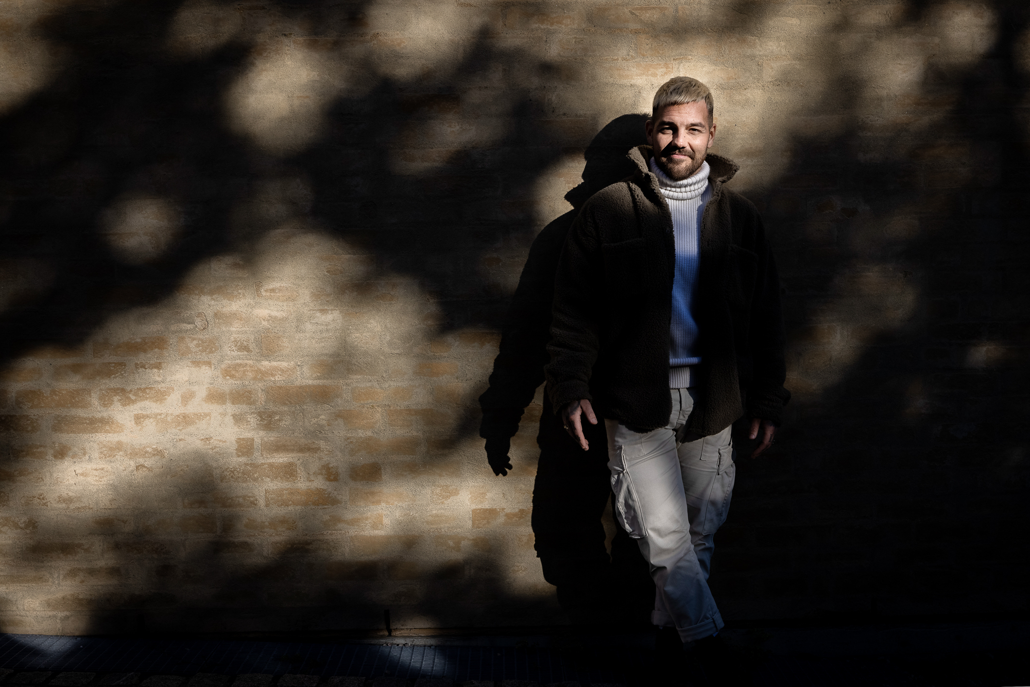 Foto: Mann i 30-årene med blondt hår og skjegg står lent mot en murvegg.