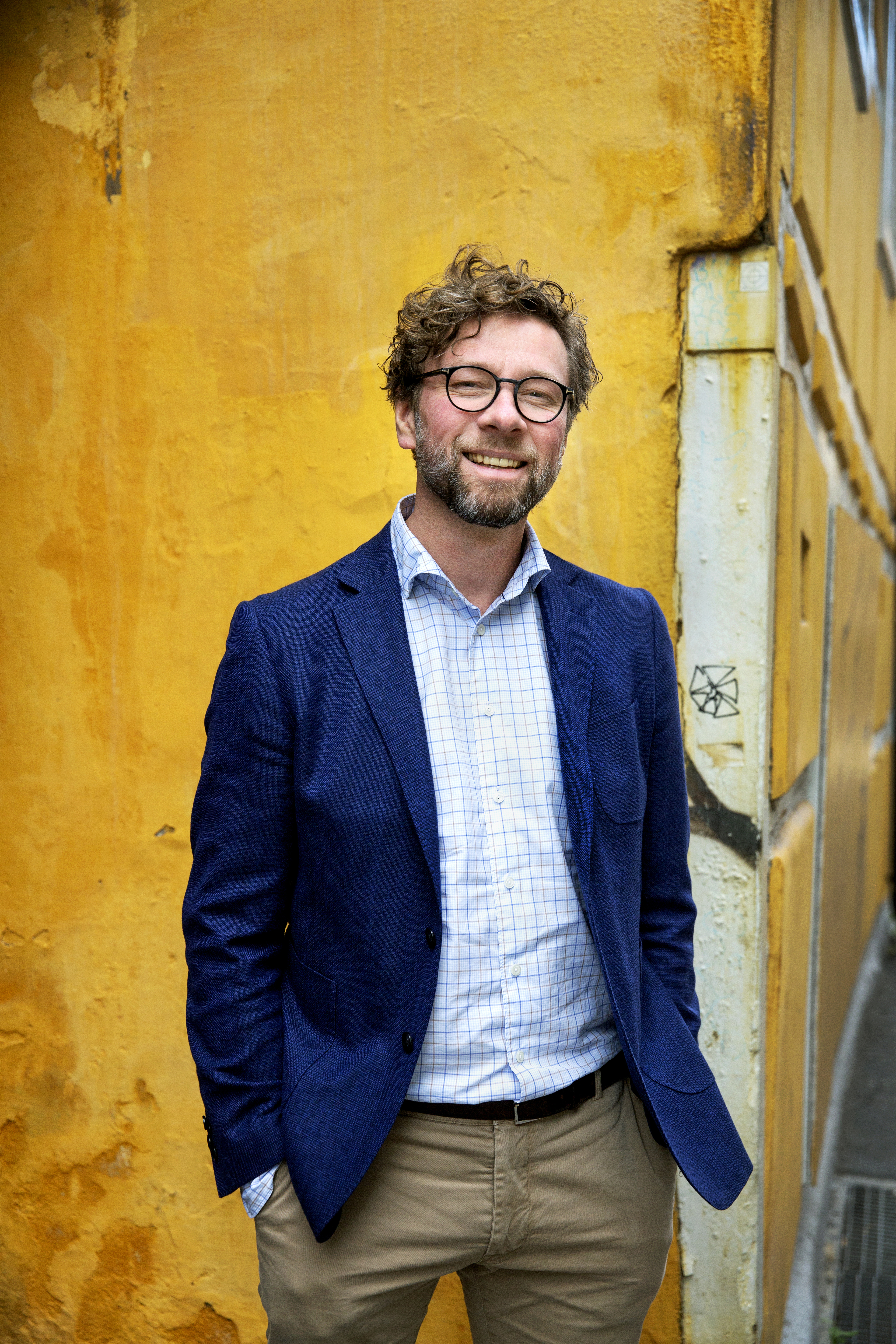 Alf Gjøsund smiler til fotografen foran en gul murvegg. Han er ikledd en beige bukse, rutete skjorte og en blå blazer.