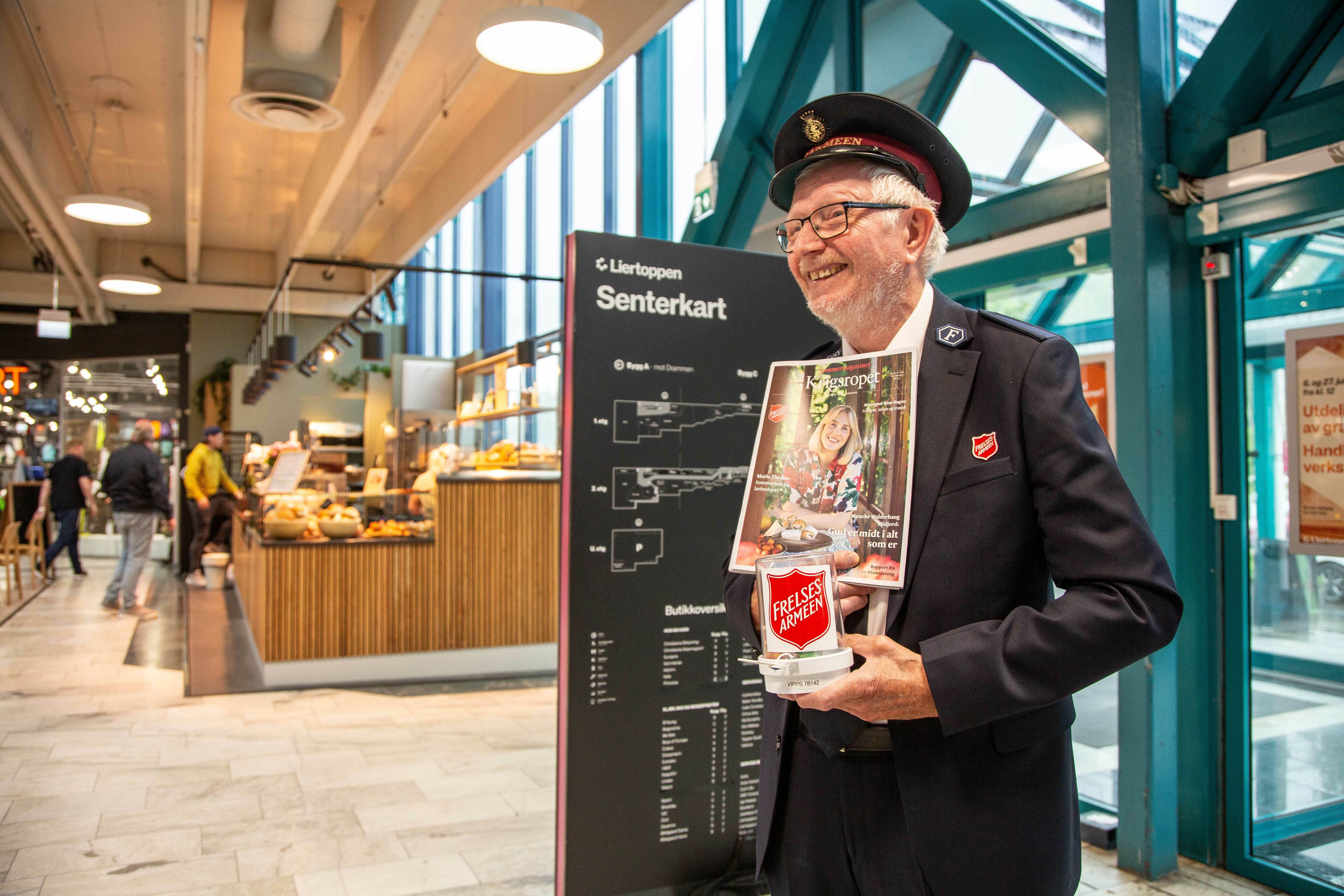 Mann med frelsesarmeuniform og lue står og selger Krigsropet rett innenfor døren til et kjøpesenter.