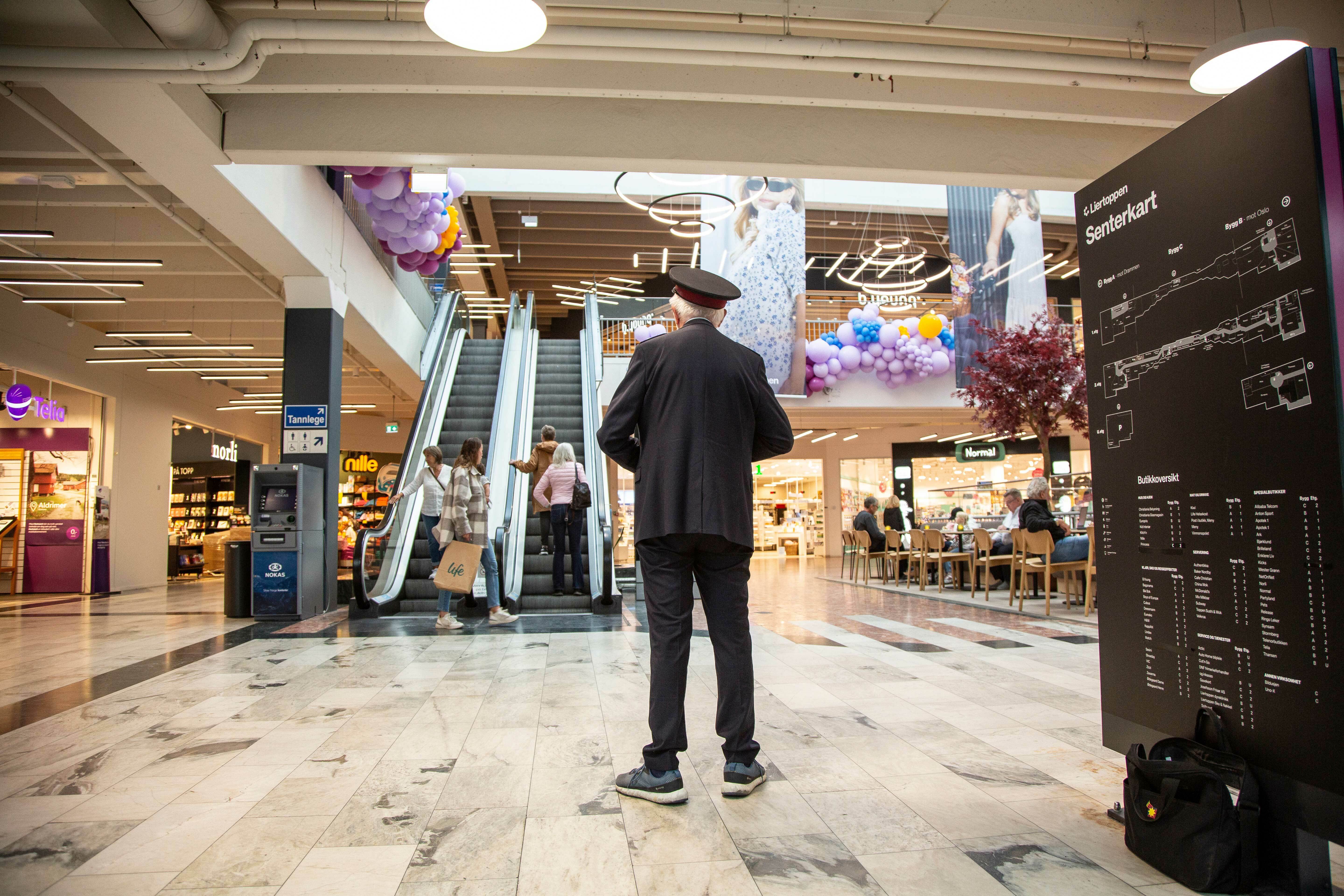 Mann med frelsesarmeuniform og lue står og selger Krigsropet på et kjøpesenter.