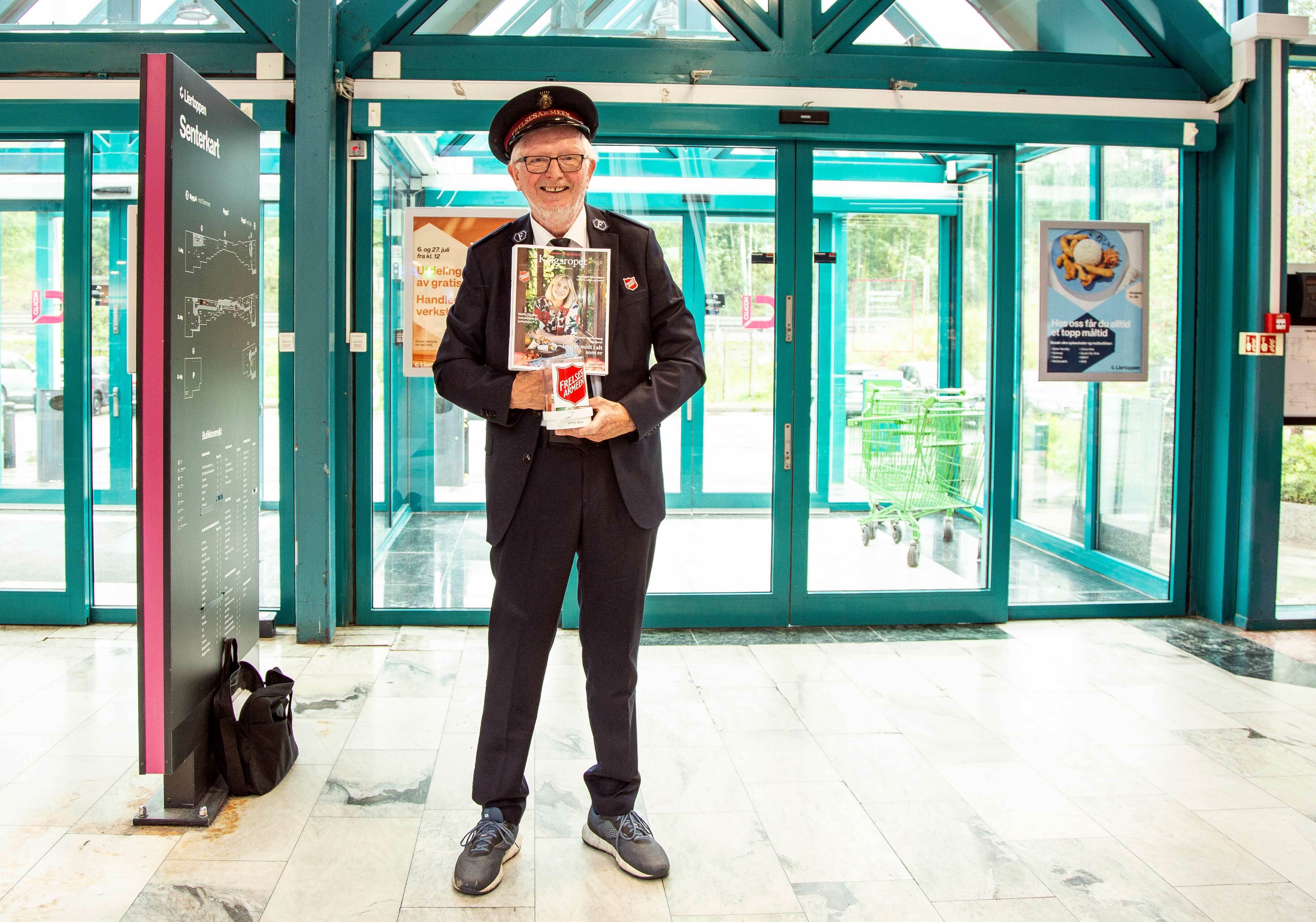 Mann med frelsesarmeuniform og lue står og selger Krigsropet rett innenfor døren til et kjøpesenter.