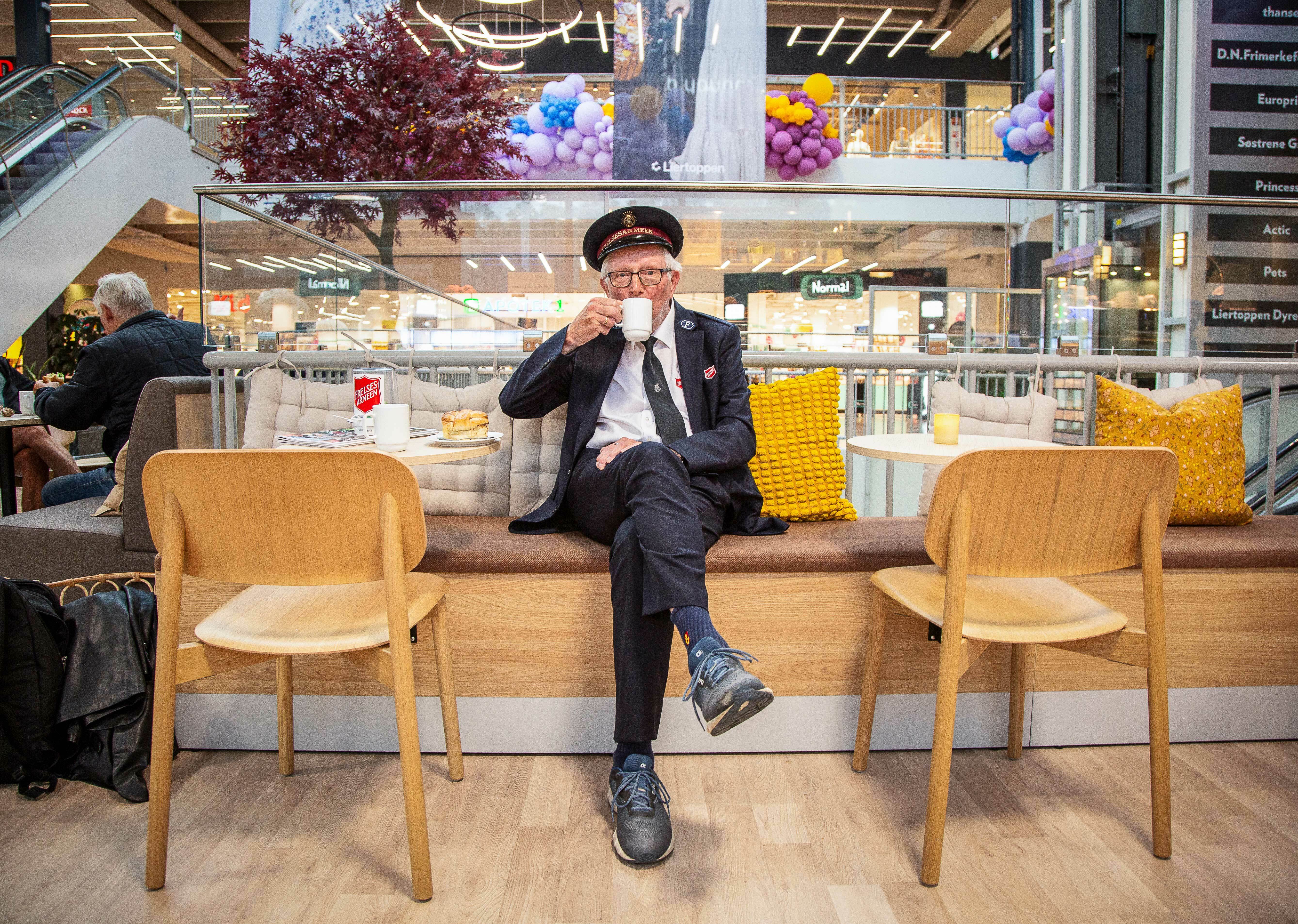 Mann med frelsesarmeuniform og lue sitter ved et kafébord og drikker kaffe på et kjøpesenter.