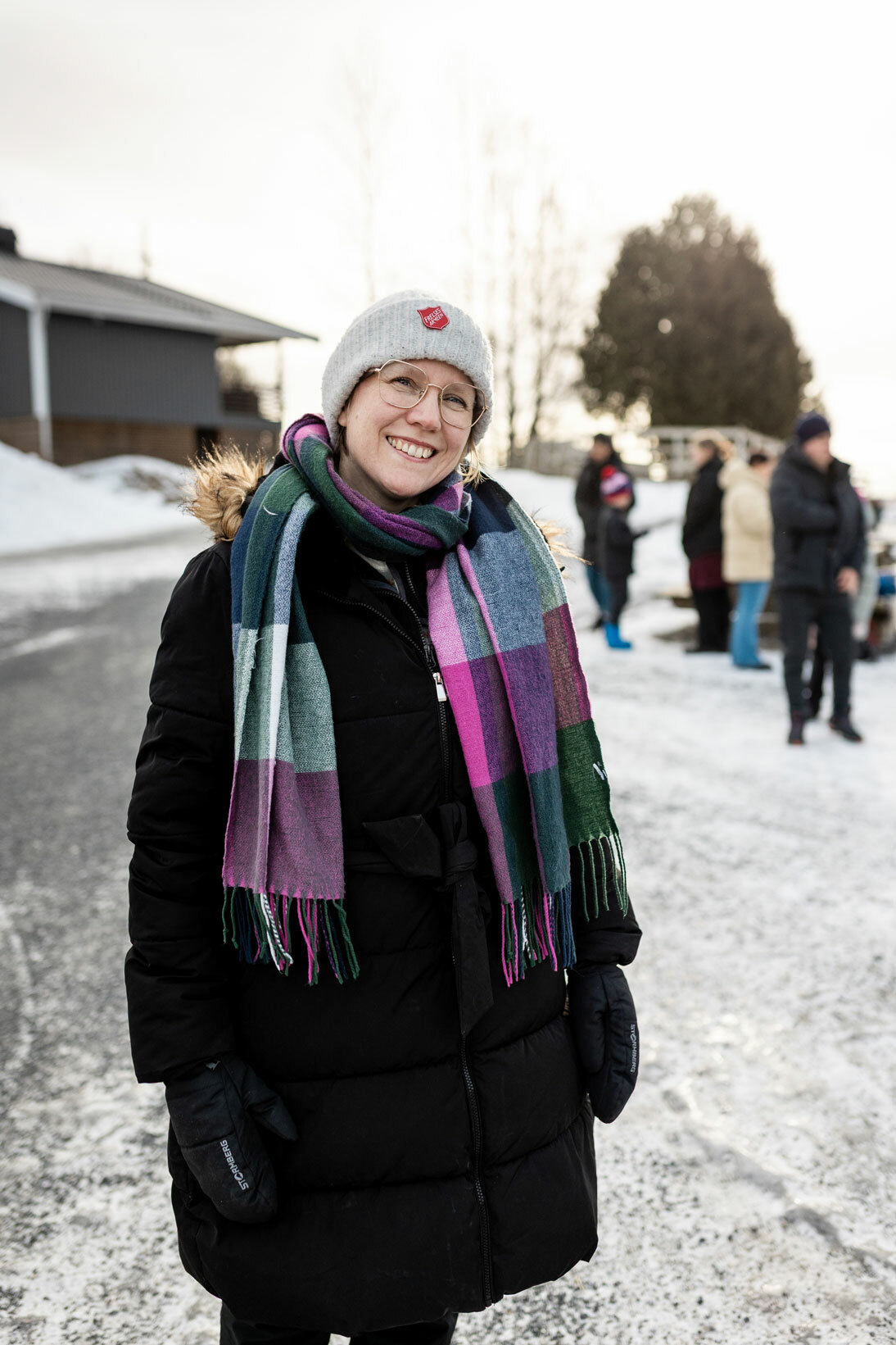 Foto: Kvinne med briller og beige lue med logo fra Frelsesarmeens står på et gårdstun med noen mennesker i bakgrunnen. Hun har på seg stor, svart boblekåpe og et rutete skjerf i rosa, burgundr og gråtoner.