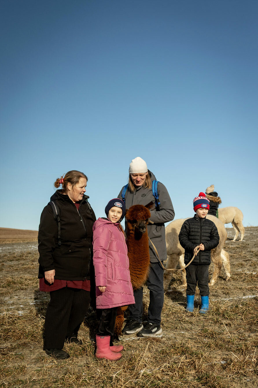 Foto: Mor, far, jente og gutt står på en eng med en alpakka i bånd. Himmelen er knall blå.