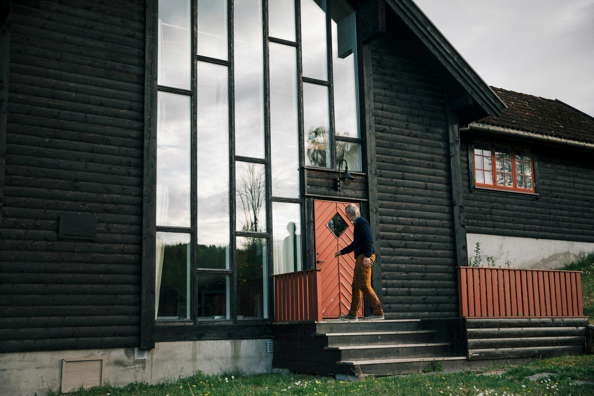 Foto: Mann som åpner en rød dør inn til et stort, brunt bygg med en hel vegg av vinduer. Bygget ser nesten ut som en moderne kirke, men med gammel inngangsdør. Den har firkantet vindu til å titte inn gjennom der vinduet er snudd 45 grader slik at spissene kommer opp, ned og ut til kantene av døra.