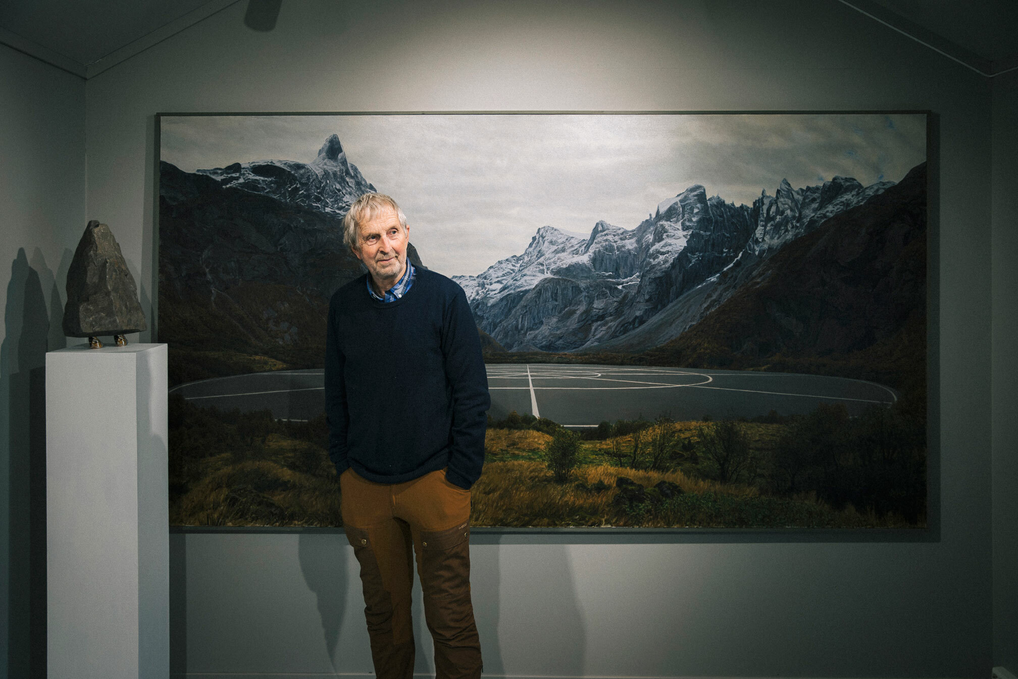 Foto: Maleri av naturtro norske fjell med hvitt dryss av snø. På fjorden under er det malt inn en flat idrettsbane med hvite markeringer der vannet skulle vært.