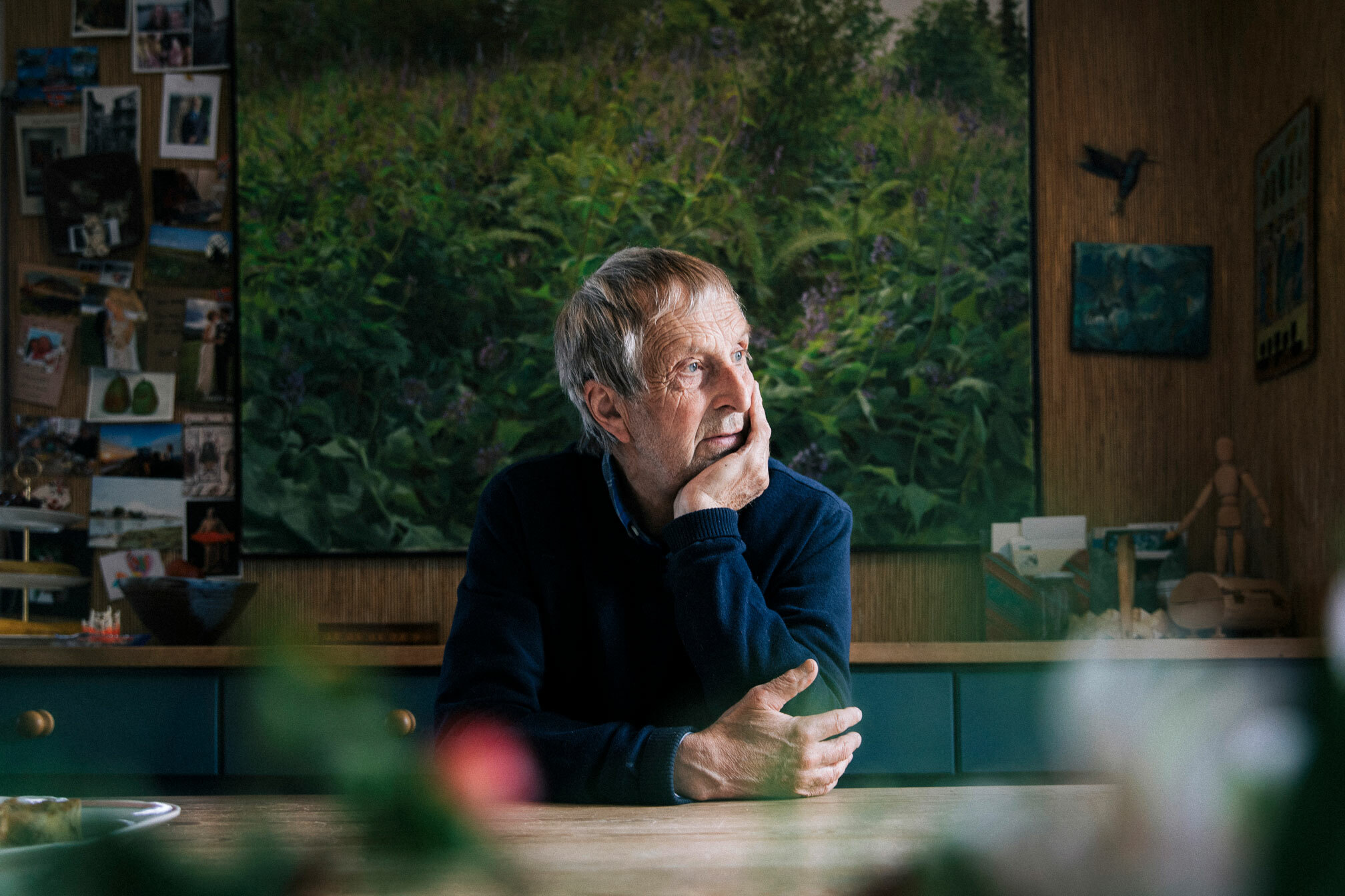 Foto: Mann sitter ved et kjøkkenbord. Han har hodet liggende i venstre hånd, den høyre ligger på bordflaten i tre. Bak ham er et stort, malt bilde av en blomstereng.