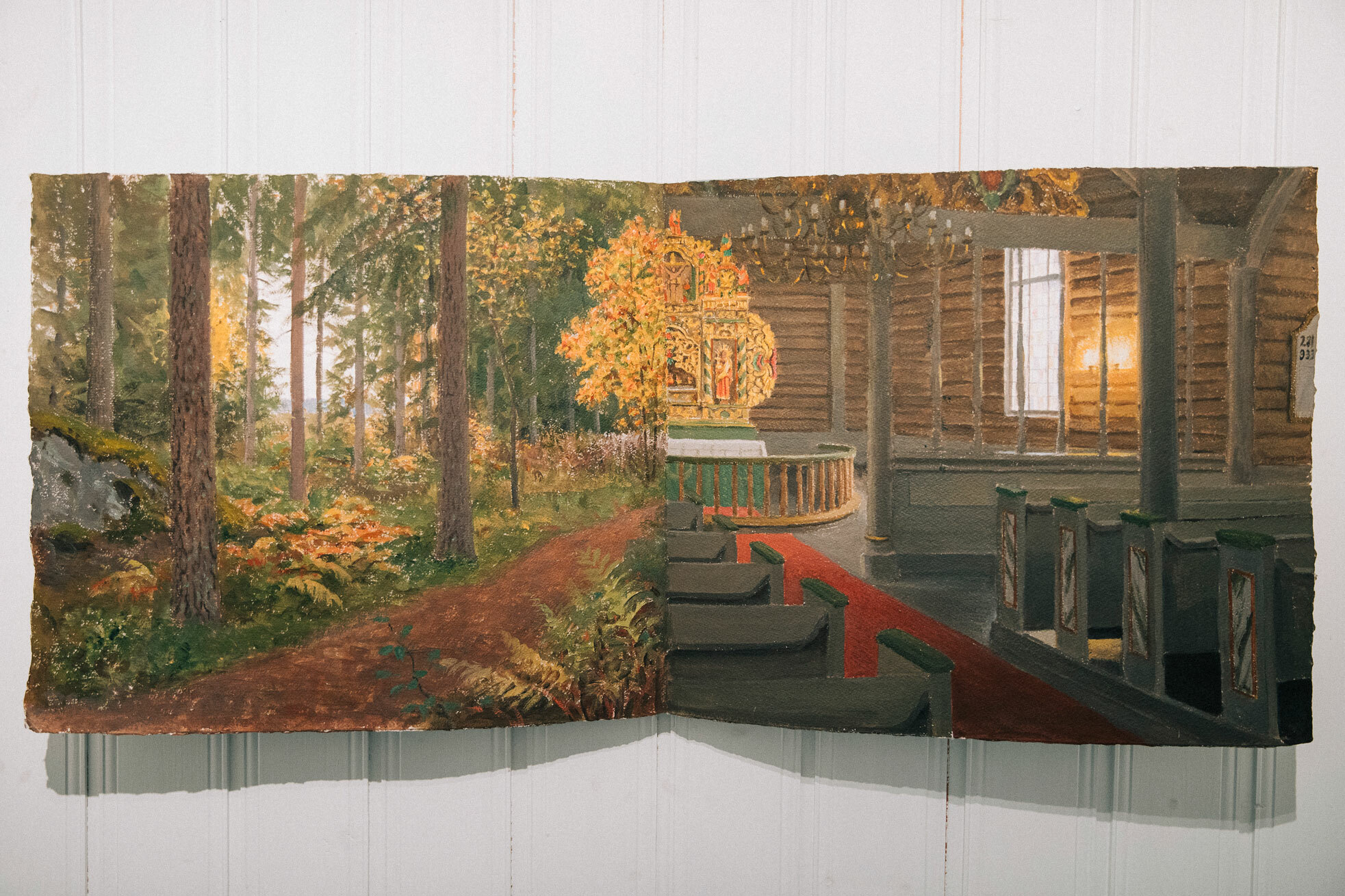 Foto: På en hvit vegg henger en akvarell på et papir som har vært brettet på midten. Delen til venstre viser naturen med trær og skogbunn og et halvt alter laget av ville blomster. Den andre halvdelen starter med alteret i gull fra Biri kirke inn mot midten, og går videre til å vise mer av kirkerommet med benkerader bakover.