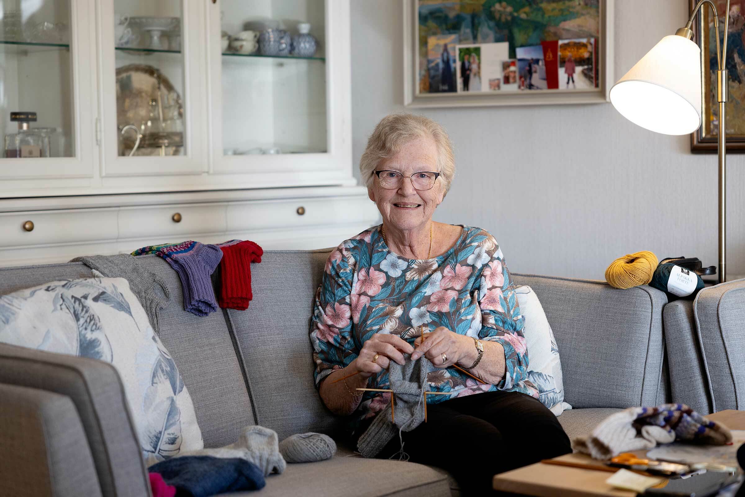 Eldre kvinne med briller og hvitt hår sitter i sofaen og strikker ullsokker.