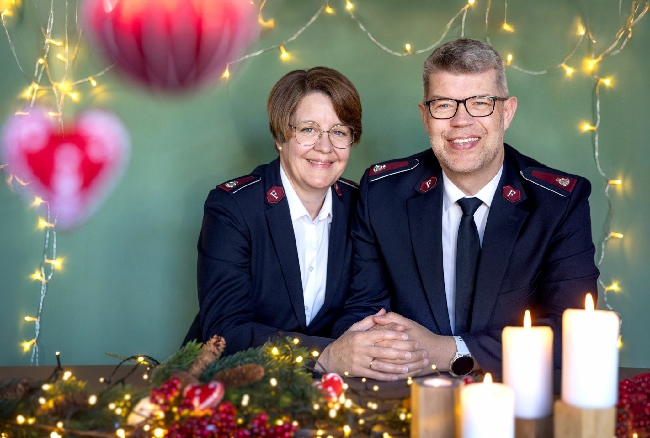 Foto: Kvinne og mann i 50-årene, begge med briller og i uniform fra Frelsesarmeen, sitter foran juledekorasjoner med grønn bakgrunn.