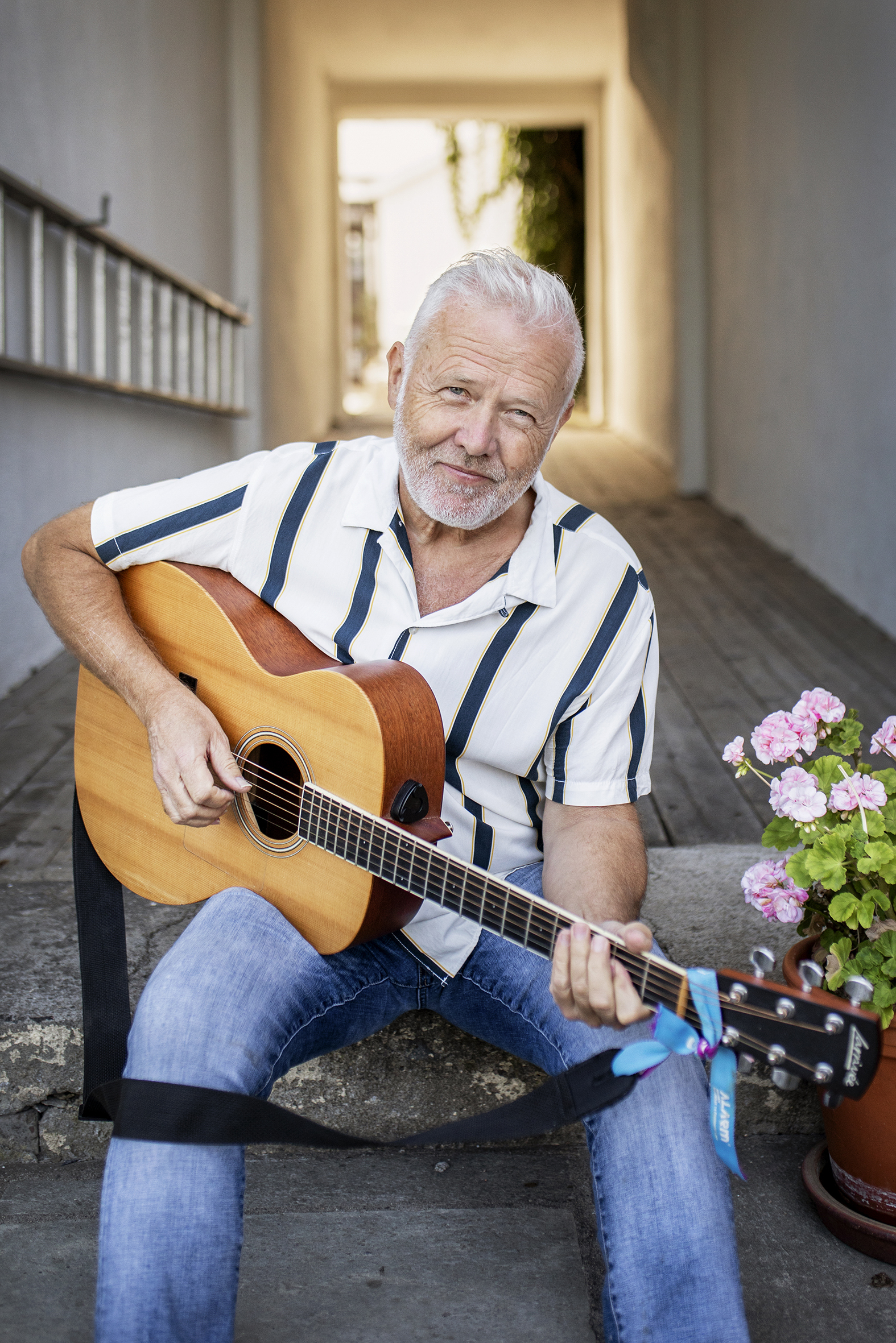 Bildet viser Amund Enger som sitter ute og spiller på en gitar. Han har på seg stripete t-skjorte og ser i kamera.