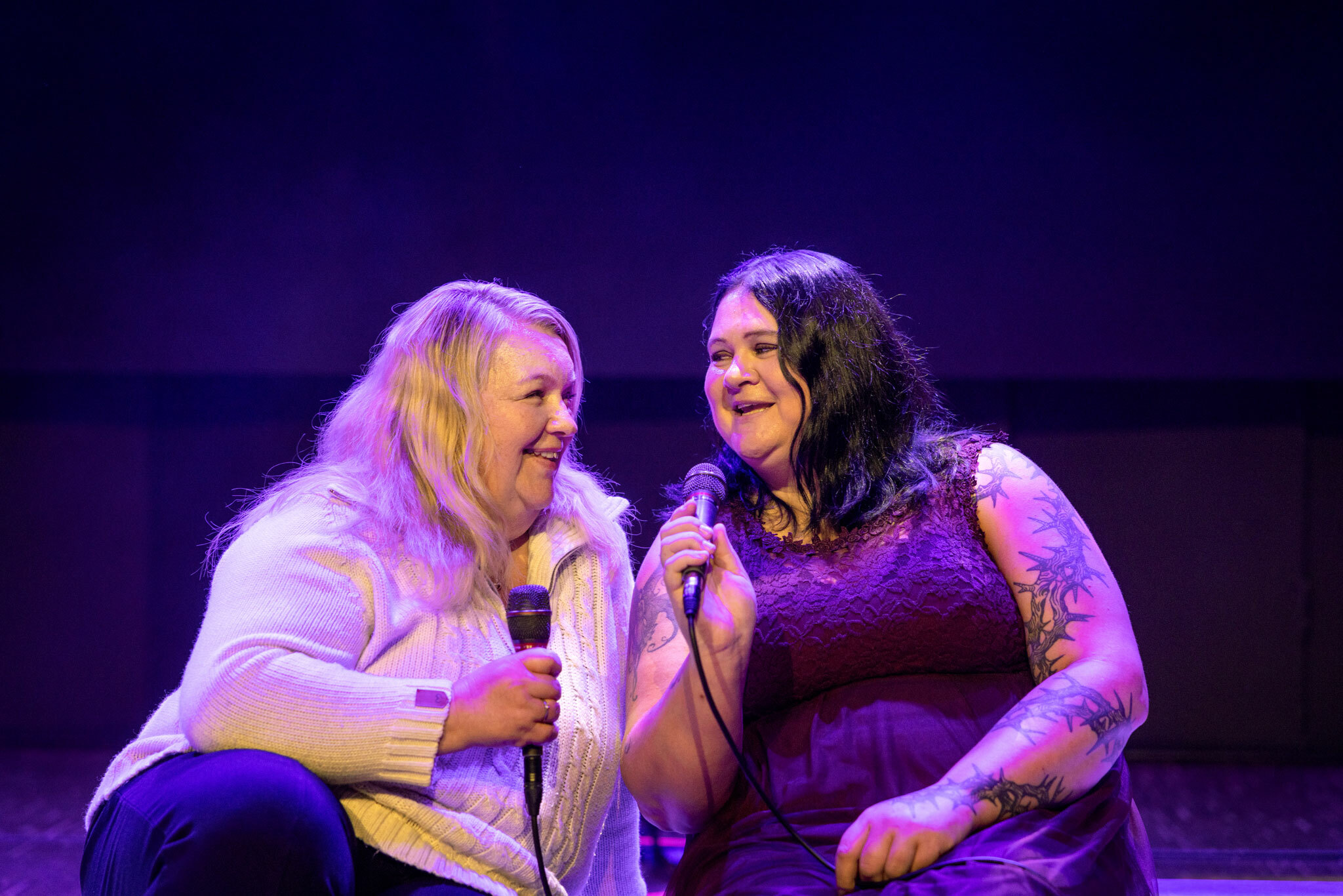 Foto: To kvinner, en lys og en mørk, holder i hver sin mikrofon. De sitter på en scenekant og lyset i rommet er blått.