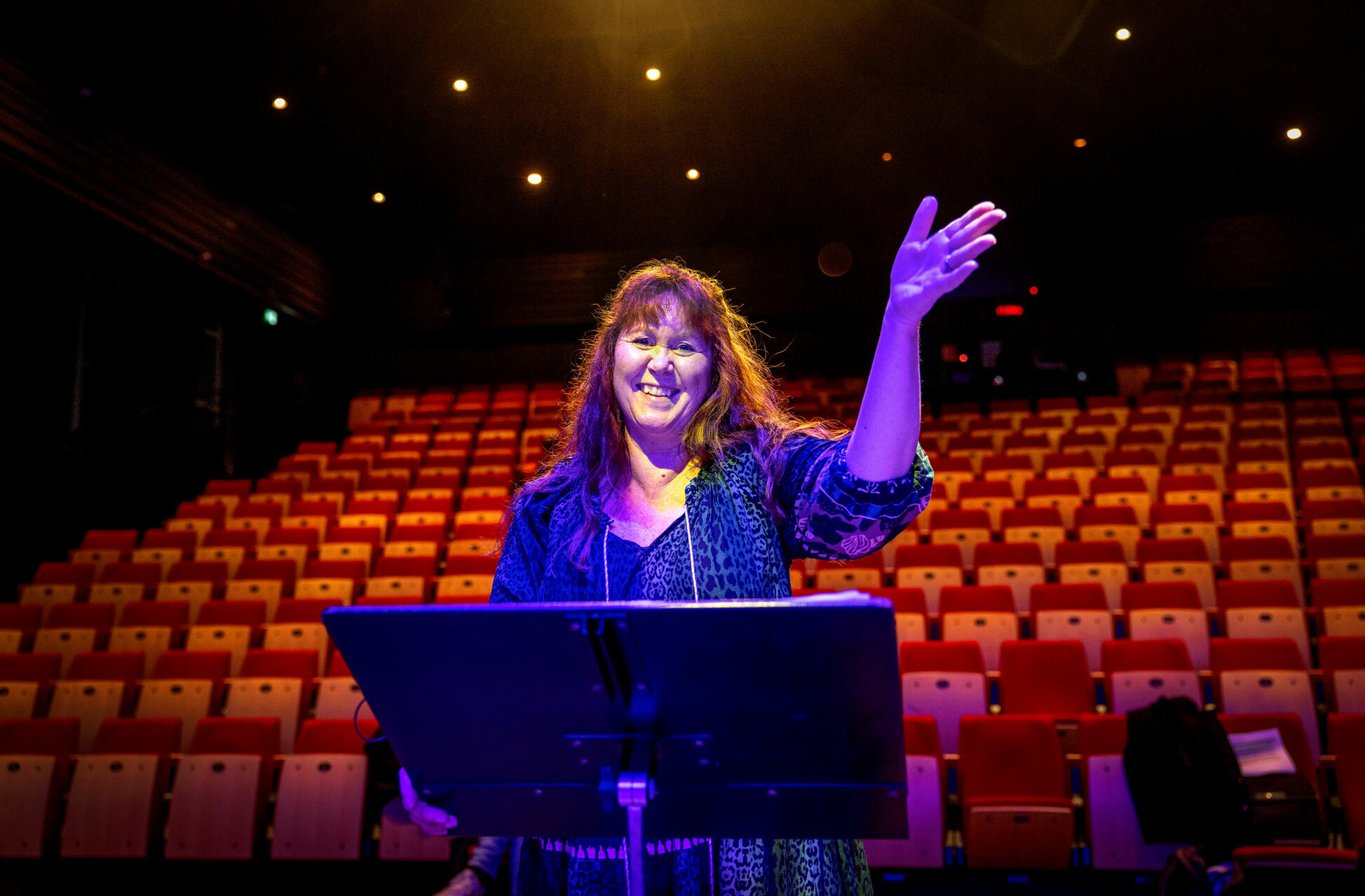 Foto: Dame som står og vinker bak et notestativ. Salen er tom for folk, men det er blått lys i en sal med rekke på rekke av røde seter.