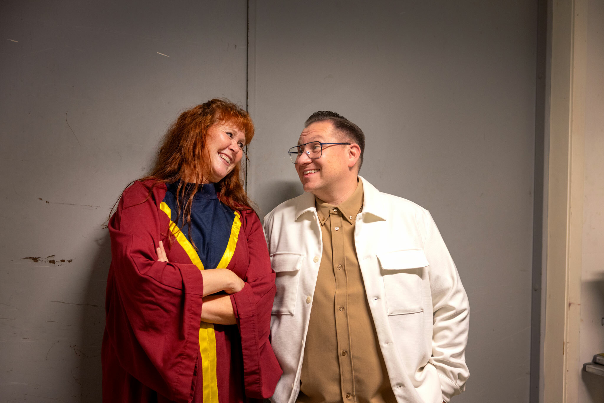 Foto: Kvinne med langt, rødlig hår i en rød korkappe står opp mot en vegge. Hun står tett sammen med en mann med briller, i beige skjorte og hvit jakke. De smiler mot hverandre.