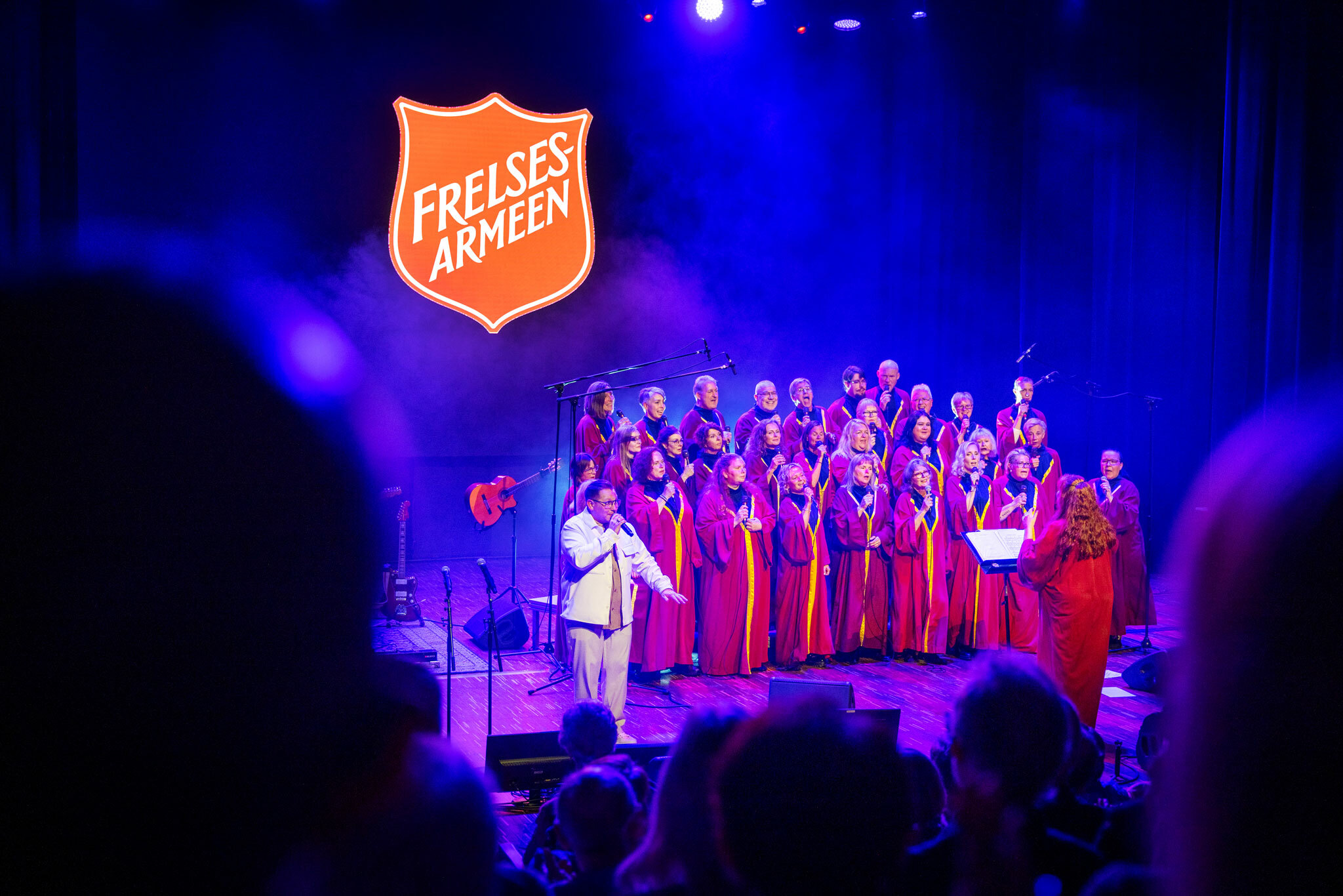 Foto: Kor i røde kapper på scene med blått lys. Til venstre står solisten.