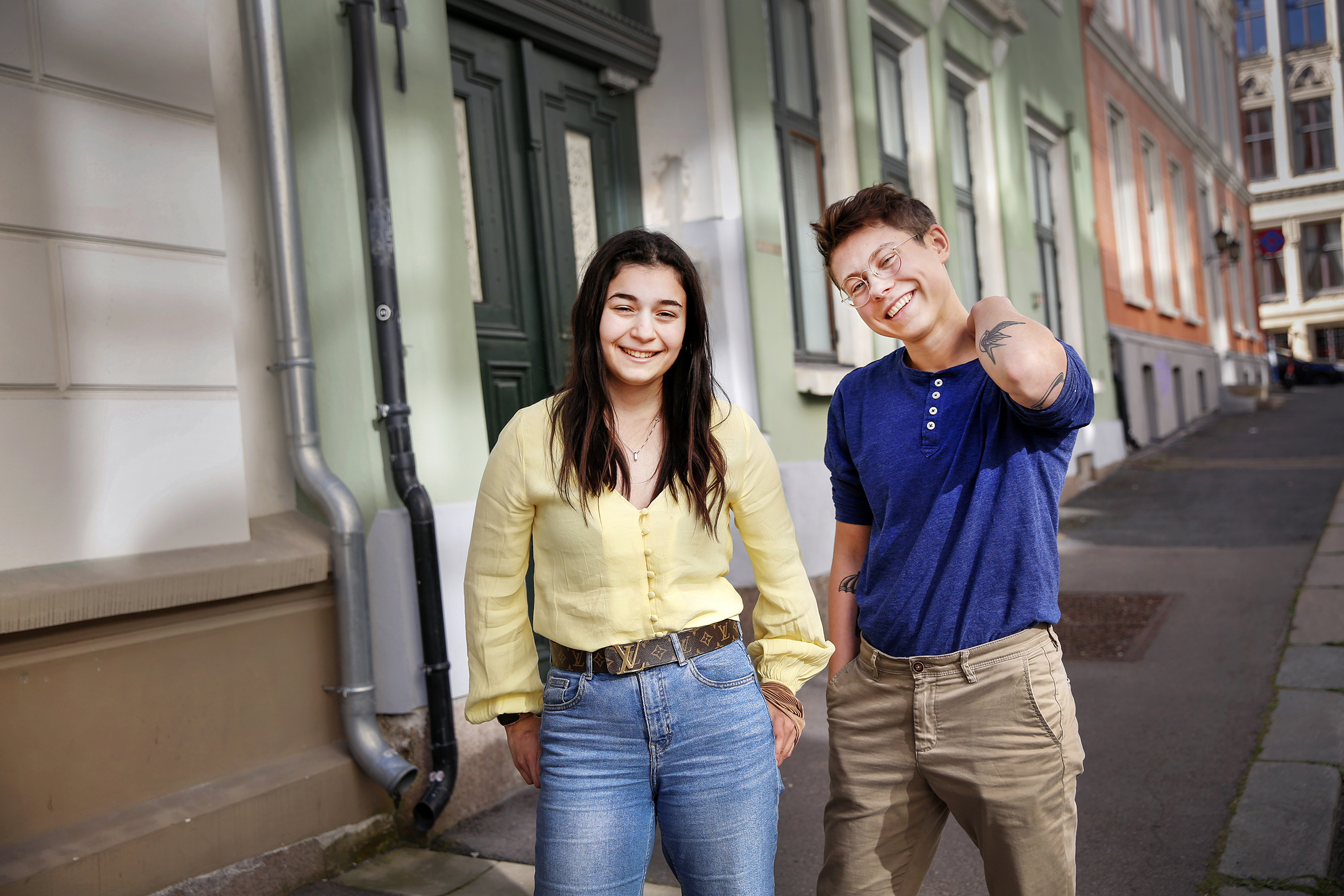 Bildet viser Dania og Leon som står i en gate med gamle bygårder. Dania har langt hår og har på seg gul skjorte og jeans, Leon har kort hår og briller og har blå t-skjorte og brune bukser. Begge smiler mot kamera.