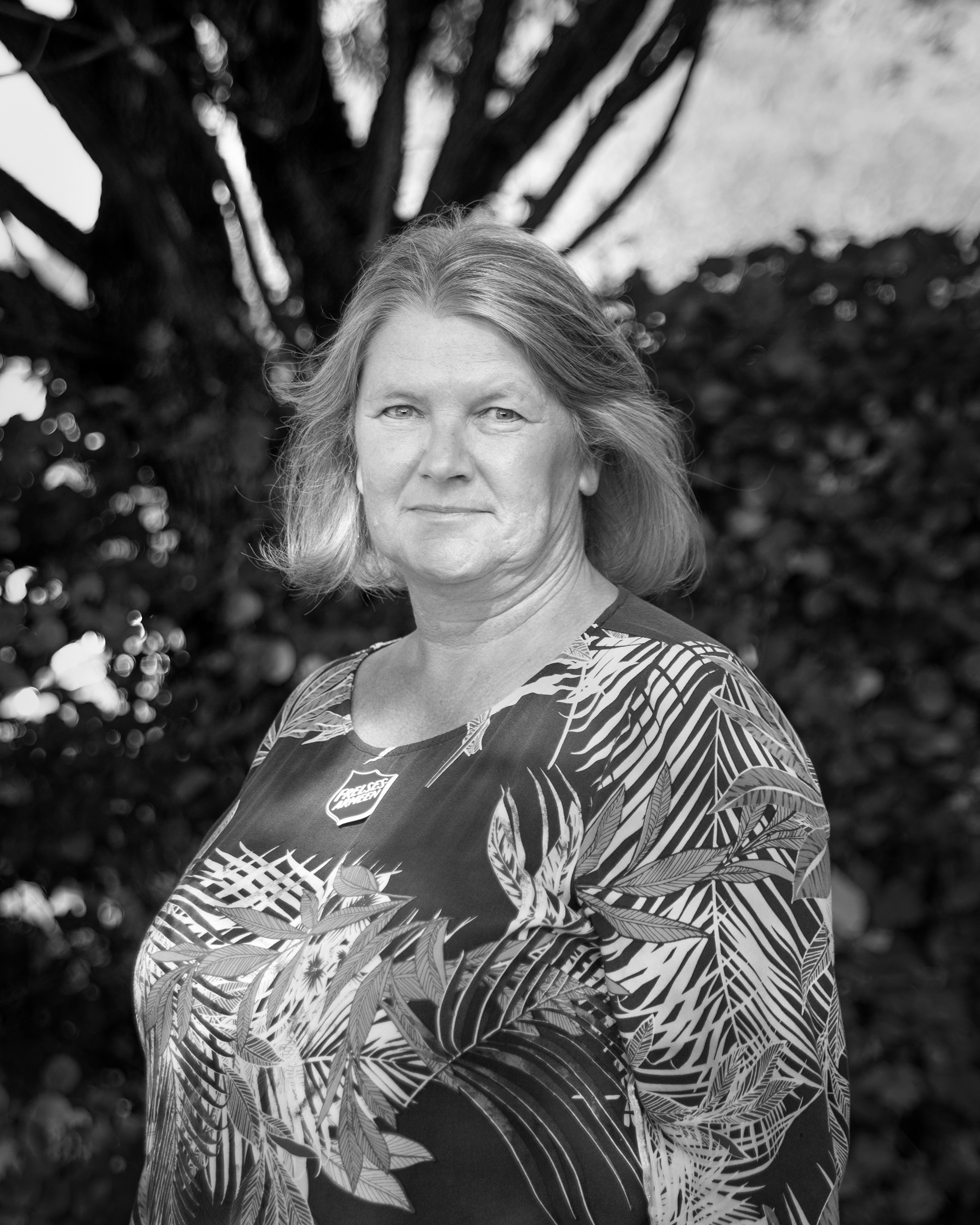 Bildet er svart-hvitt og viser en kvinne med halvlangt hår som står utendørs foran noen busker. Hun har på seg en mønstrete genser.