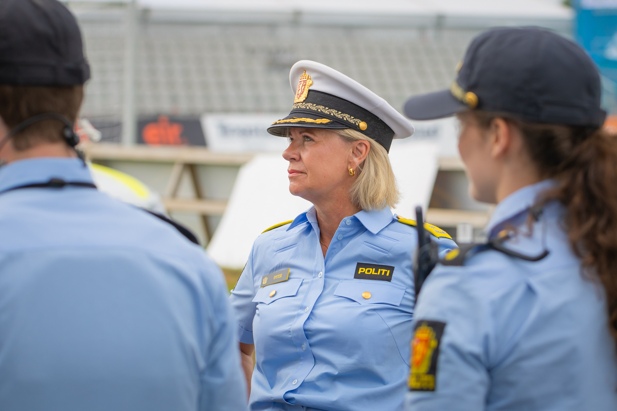 Tre politi i uniform med blå skjorter står sammen med stadion-reklame og tribune i bakgrunnen. To med ryggen til. To kvinner og en mann. Politikvinnen til høyre har mørke krøller i hestehale og mannen  til venstre  er mørk blond og kortklipt. I midten står en middelaldrende kvinne med lyst hår og politimesterlue som er hvit med sort skygge.