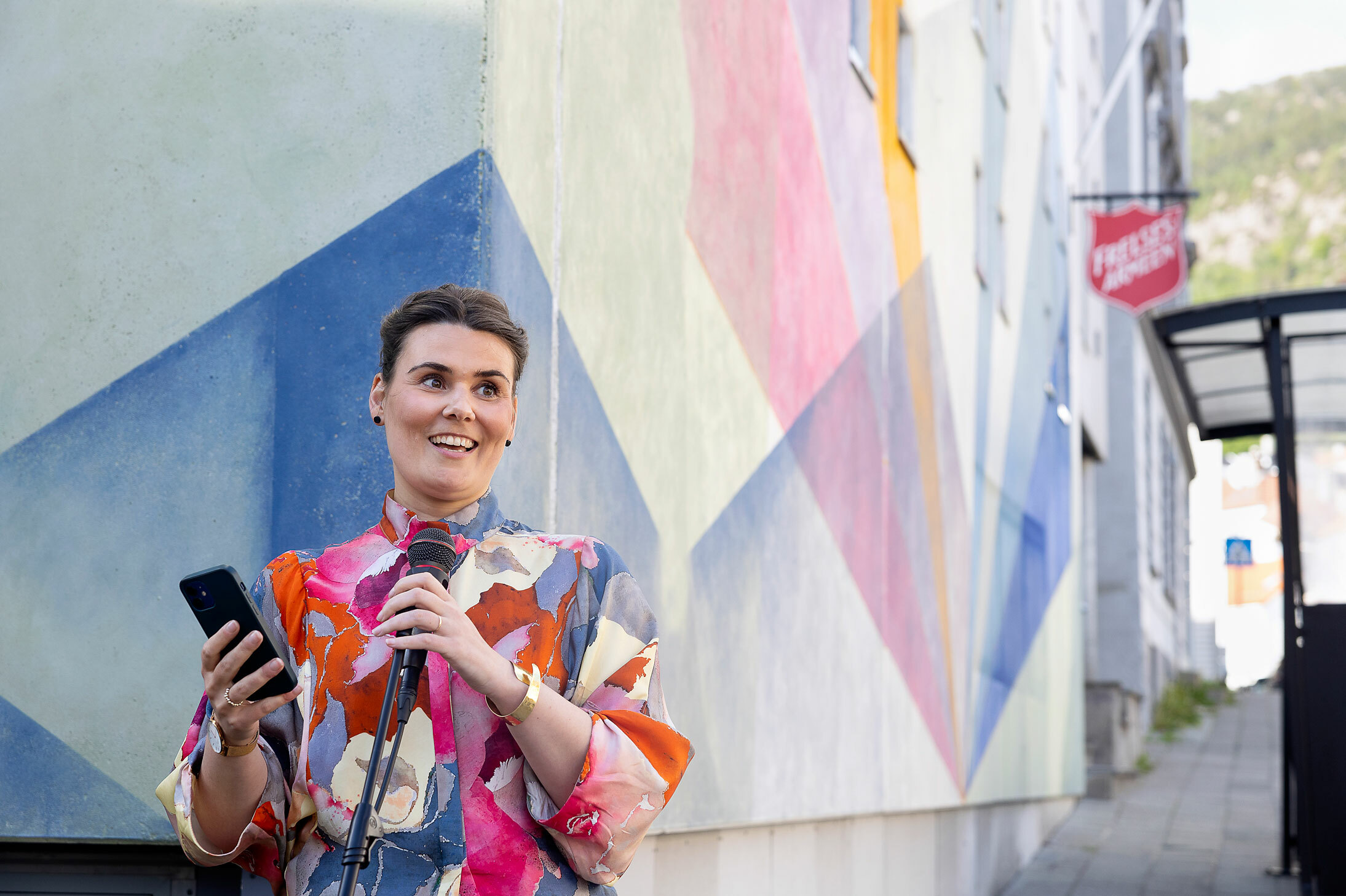 Foto: Kvinne i 30-årene snakker i en mikrofon. Hun har notatene på mobilen som hun holder i den andre hånden. I bakgrunnen er det en fargerik murvegg og skiltet til Frelsesarmeen.