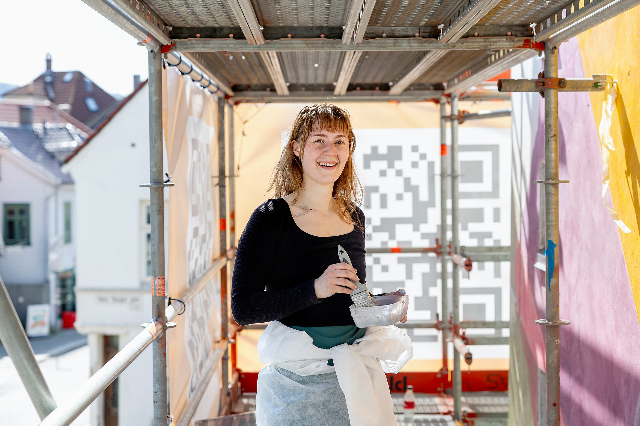 Foto: Smilende ung jente står høyt oppe i et stilas med en malerkost og en plastikkskål med maling i hendene. På murveggen til høyre har hun malt abstrakte former i gult og lilla.