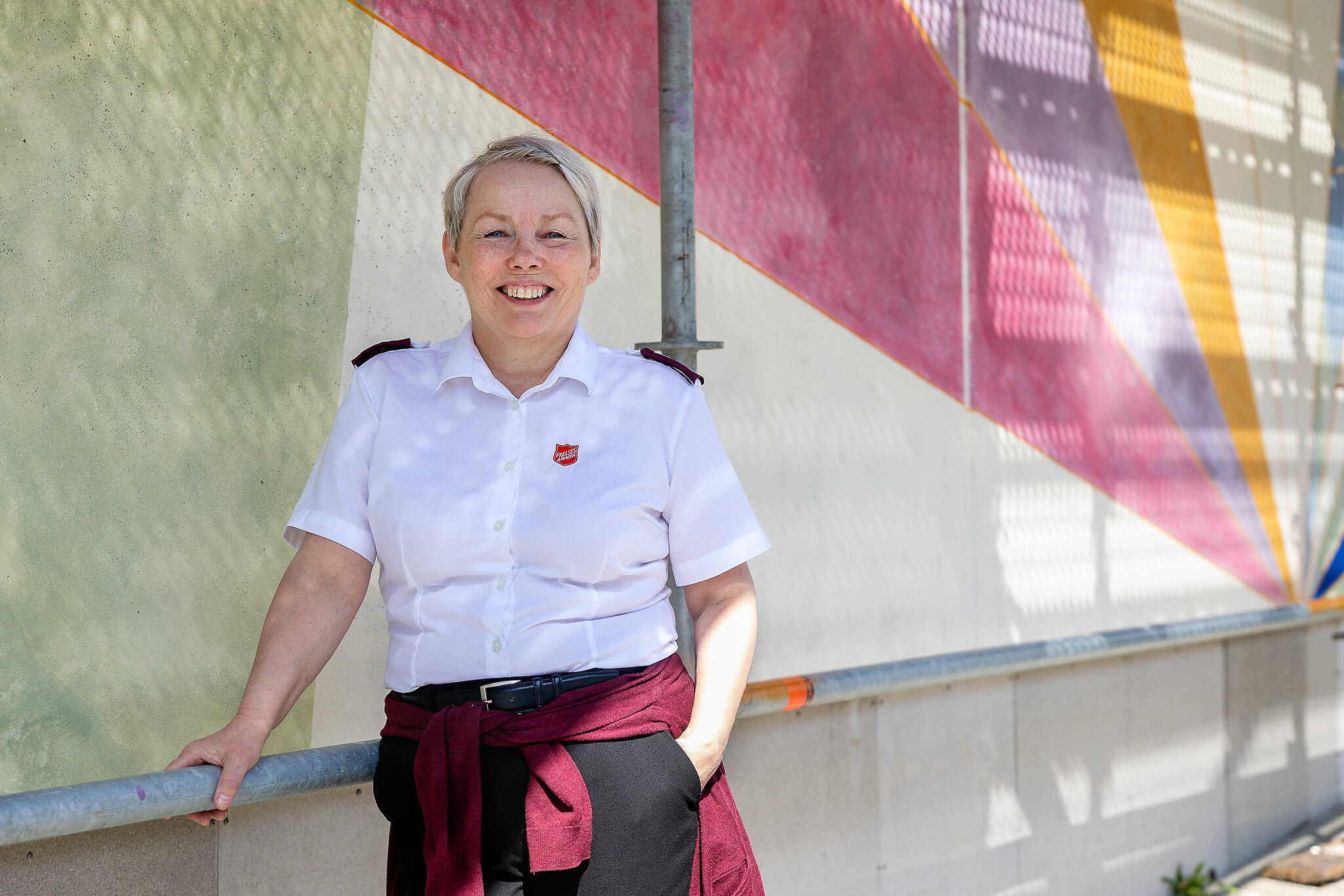Foto: Kvinne med kort hår og hvit uniformsskjorte fra Frelsesarmeen står ved veggen. Hun står på bakkeplan, men rundt og over henne står stilaset.