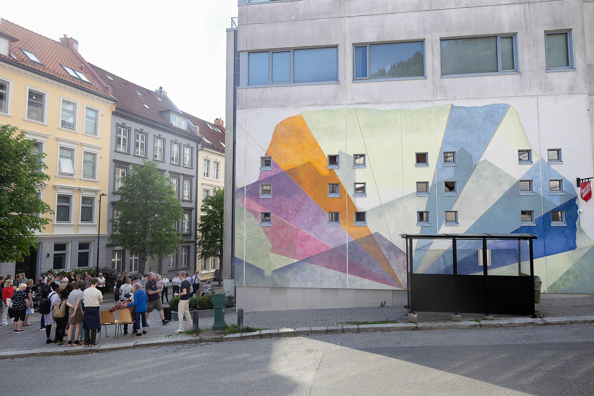 Foto: Fargerik murvegg på et moderne betongbygg ses i sammenheng med resten av gata med sine gamle bygårder fra slutten av 1800-tallet. I gata midt i mellom står en menneskemengde.