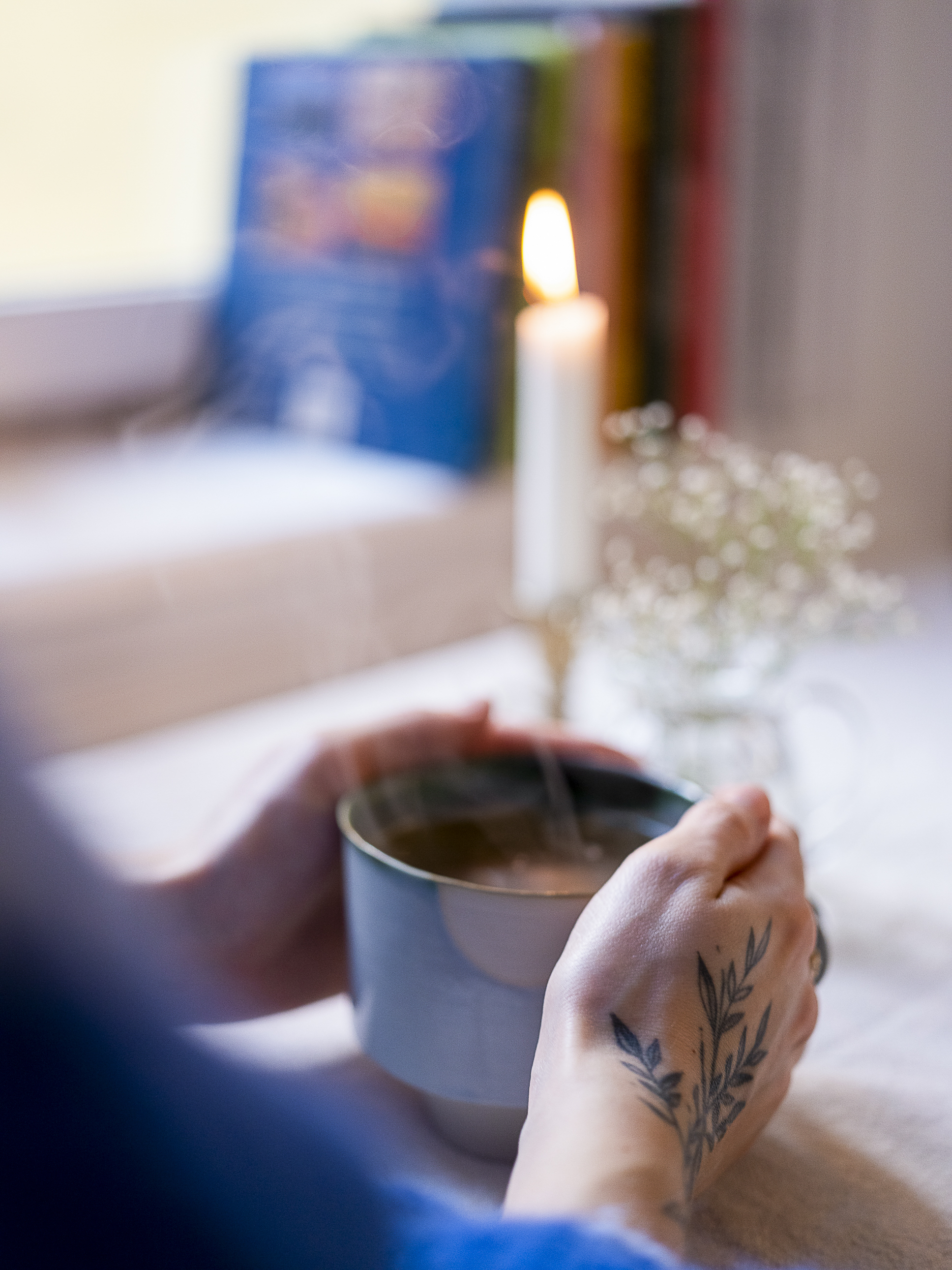 To hender holder en rykende varm kopp med te. På bordet står det et tent lys og blomsterslør i en liten vase.