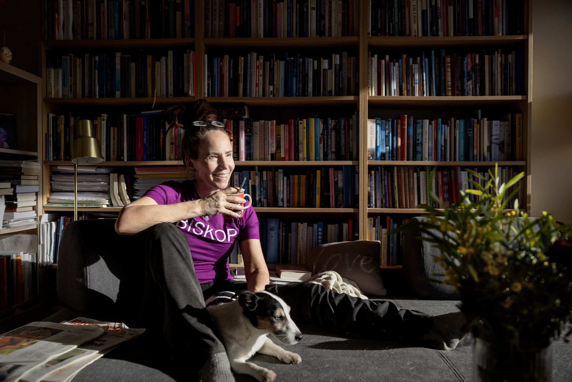 Foto: Kvinne i lilla T-skjorte med ordet BISKOP på sitter på en sofa sammen med en hvit hund med svarte og beige tegninger. I hånden holder hun en tekopp med Lille My på og bak henne er det hyllemeter med bøker.