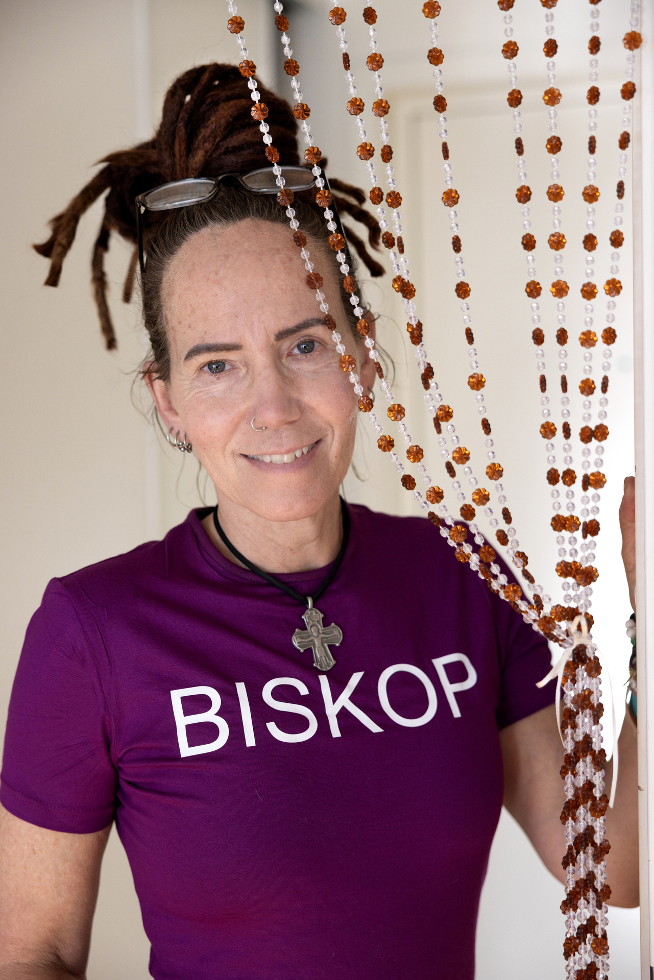 Foto: Kvinne kommer fram bak et forheng laget av brune og hvite perler. Hun har rastahår som er satt opp i en dult og har lesebriller oppå hodet. Hun har på seg en lilla T-skjorte hvor det står biskop med store bokstaver. Smykket i halsen er et stort sølvfarget kors i et sort, kort kjede.
