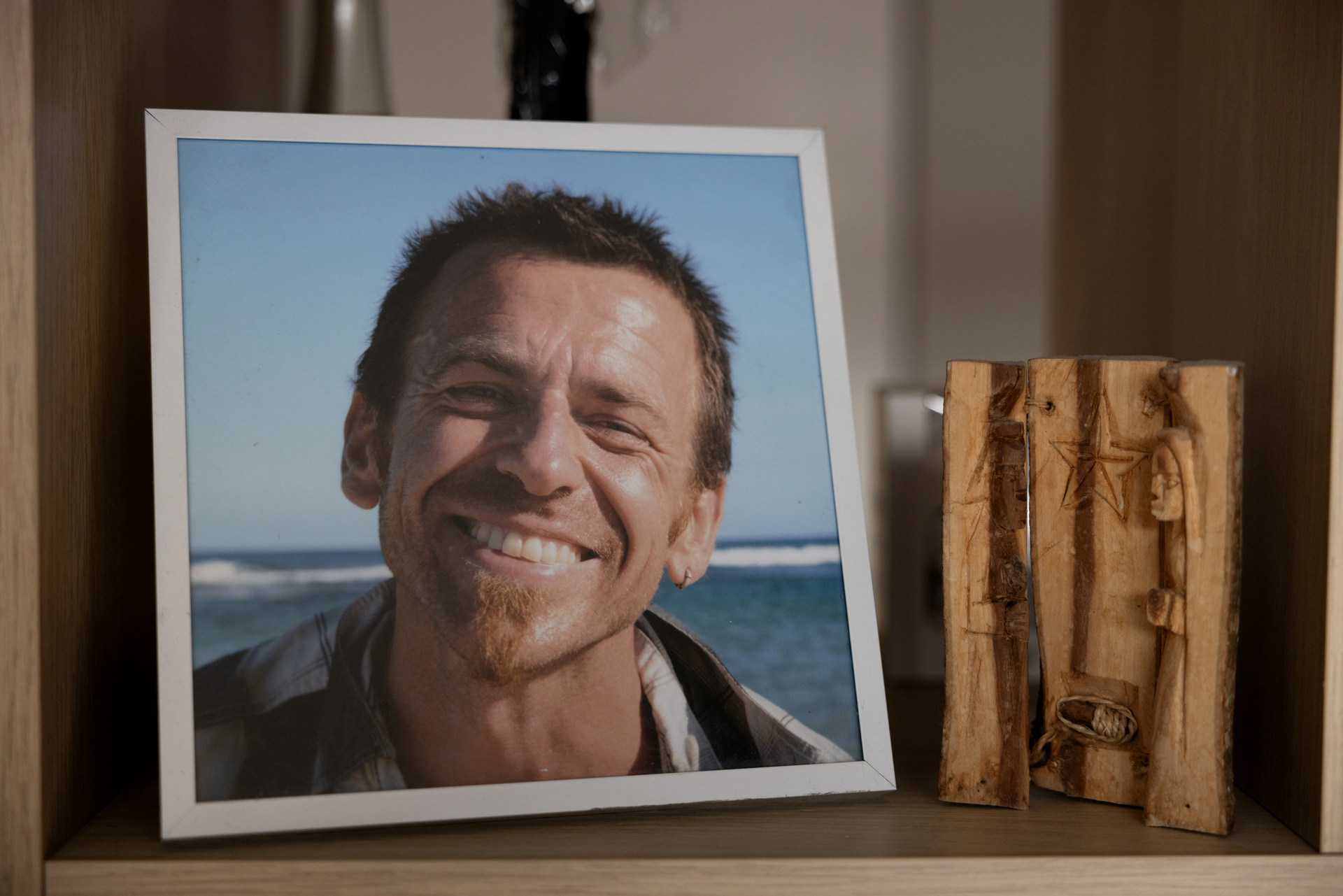 Foto: I en ramme fra hylla smiler en mann med mørkt hår fra et fotografi. I bakgrunnen er det blå himmel og hav.