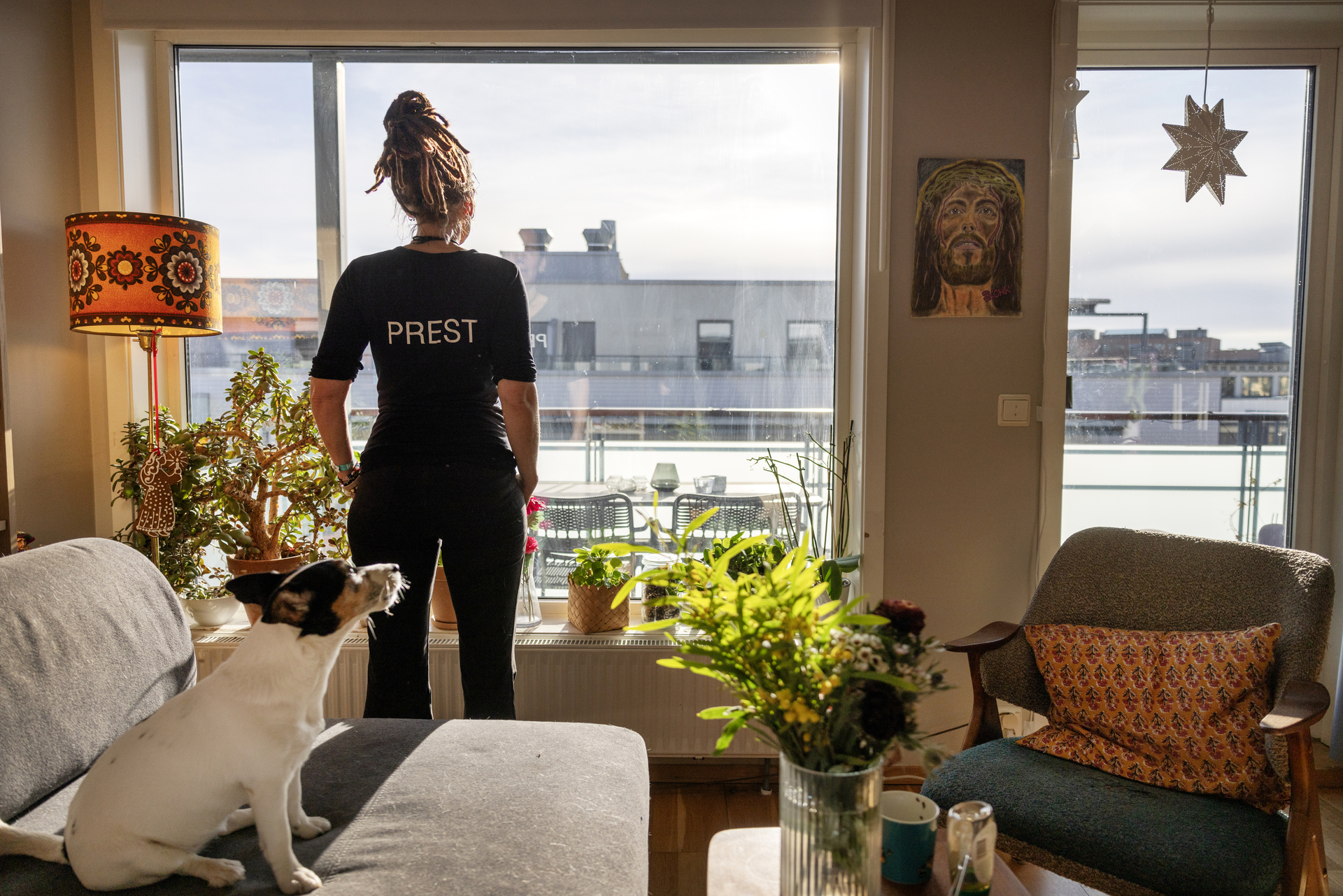 Foto: Kvinne i svar T-skjorte med ordet prest i store bokstaver på ryggen. Hun ser ut av et stor stuevindu, på høye bygninger i Oslo. På sofaen sitter en liten hund.
