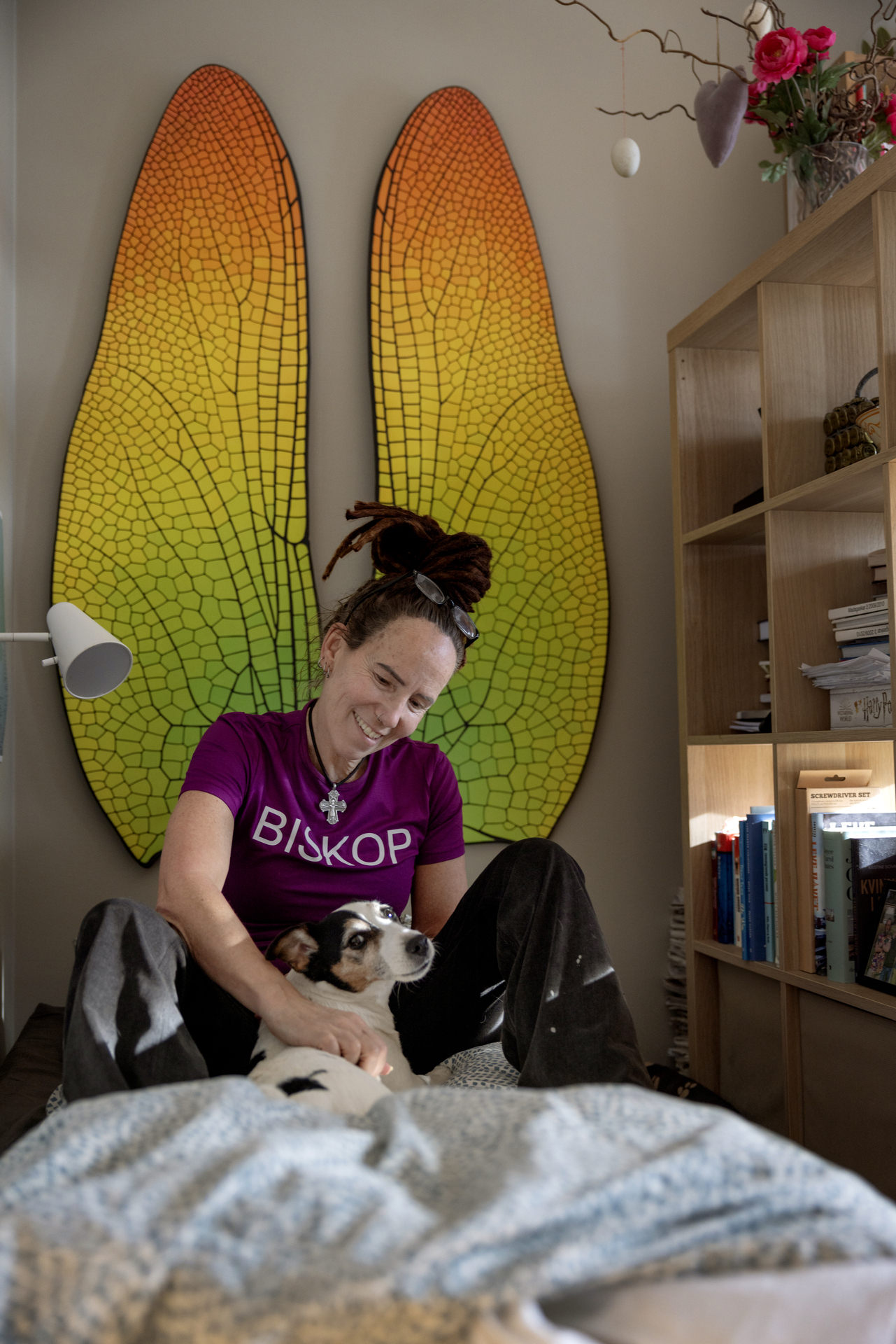 Foto: Kvinne sitter på en madrass på gulvet og klapper en hund. Bak på veggen henger et par gigantiske vinger.