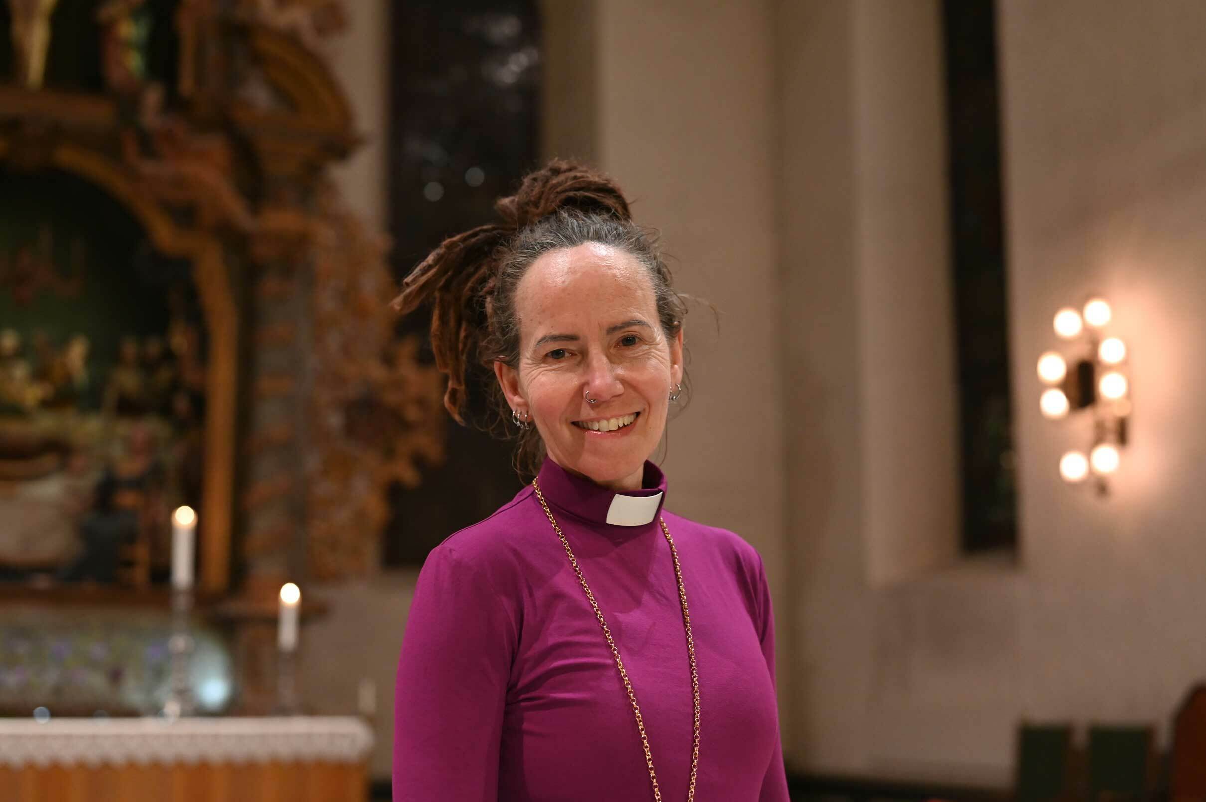 Foto: Kvinne med oppsatt rastahår smiler mot kamera. Hun har på seg lilla prestedrakt med hvit snipp og er fotografert i Oslo Domkirke.