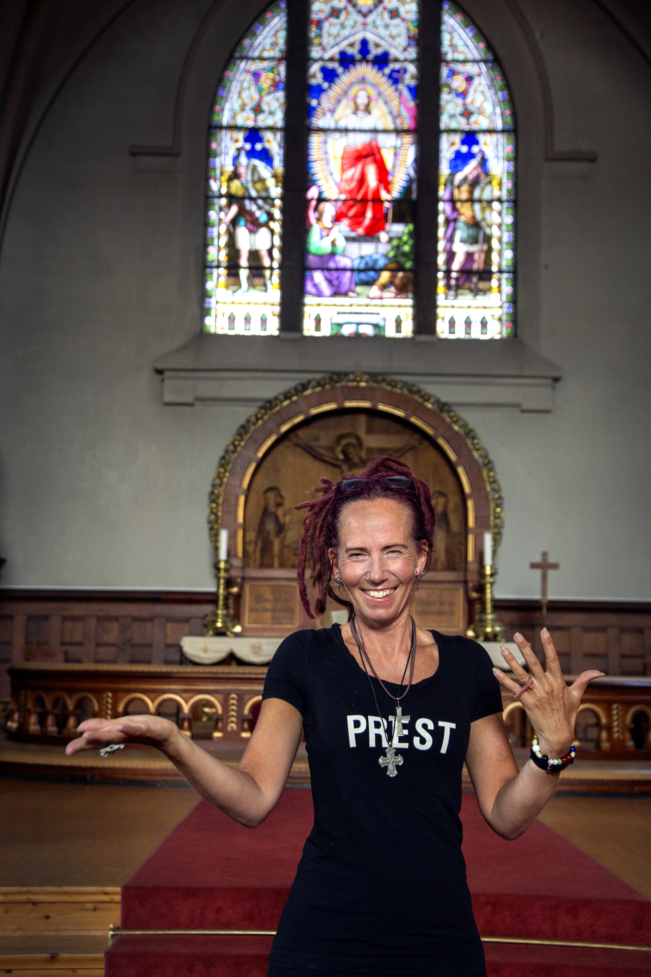 Foto: Kvinne med svart T-skjorte der det står PREST holder ut armene i et kirkerom. Hun har rastafletter og et stort kors i søv rundt halsen.
