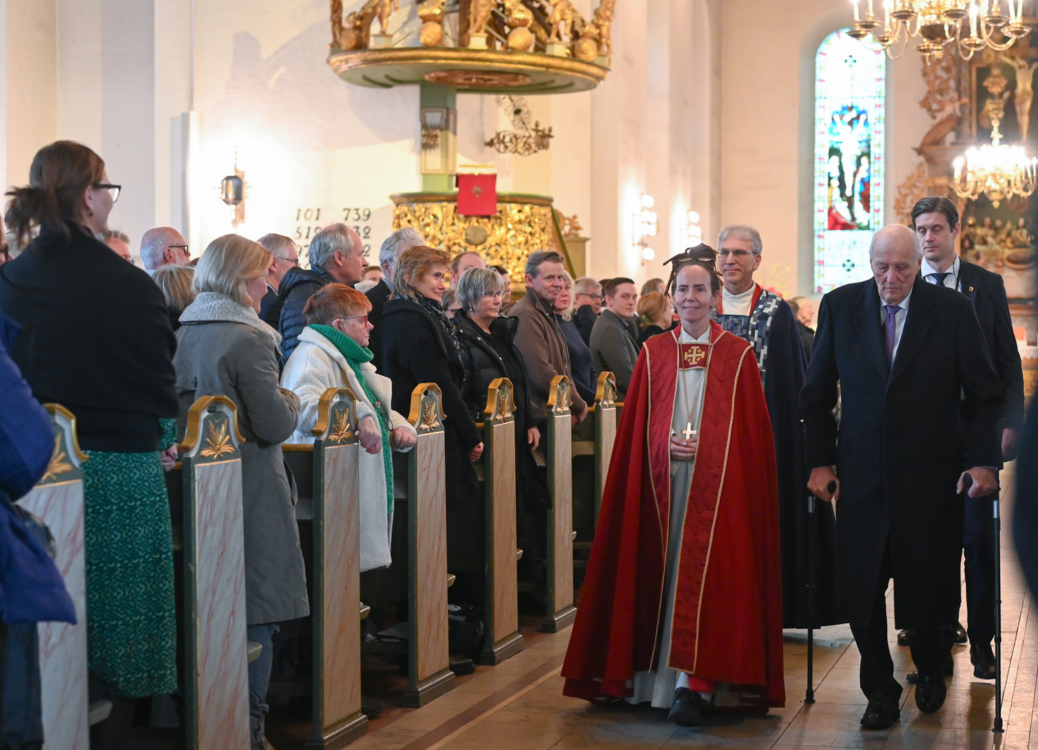 Foto: Biskopen i kappe og kongen går sammen ut av kirken med flere folk bak seg. Folk står i benkeradene.