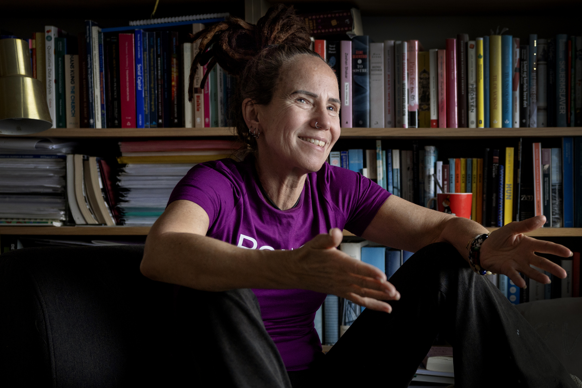 Foto: Kvinne i lilla T-skjorte sitter foran en bokhylle. Hun snakker mens hun gestikulerer med armene.