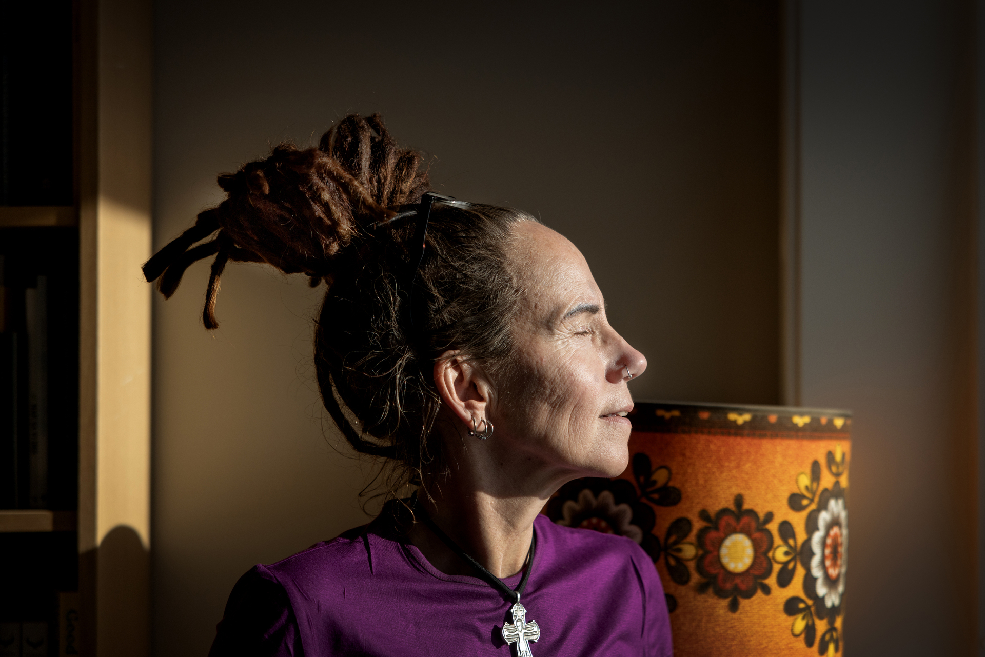 Foto: Kvinne med lukkede øyne ser mot sollyset i rommet. Hun har et kors rundt halsen og bak sees en oransje retro lampeskjerm.