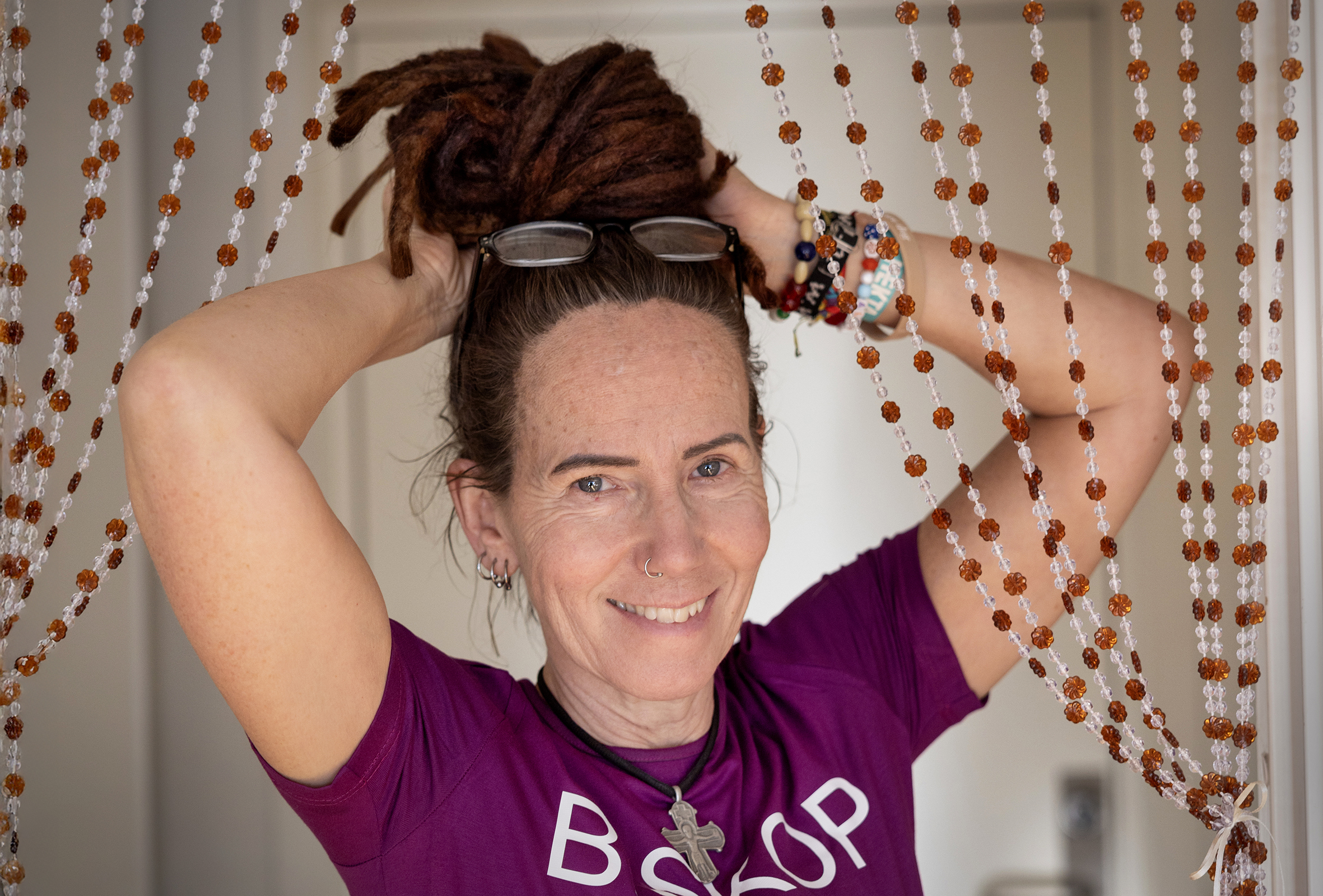 Foto: Kvinne kommer fram bak et forheng laget av brune og hvite perler. Hun har rastahår som hun setter opp i en dult med begge hendene. På hodet sitter et par lesebriller. Hun har på seg en lilla T-skjorte hvor det står biskop med store bokstaver. Smykket i halsen er et stort sølvfarget kors i et sort, kort kjede. Rundt venstre håndledd har hun mange fargerike armbånd.