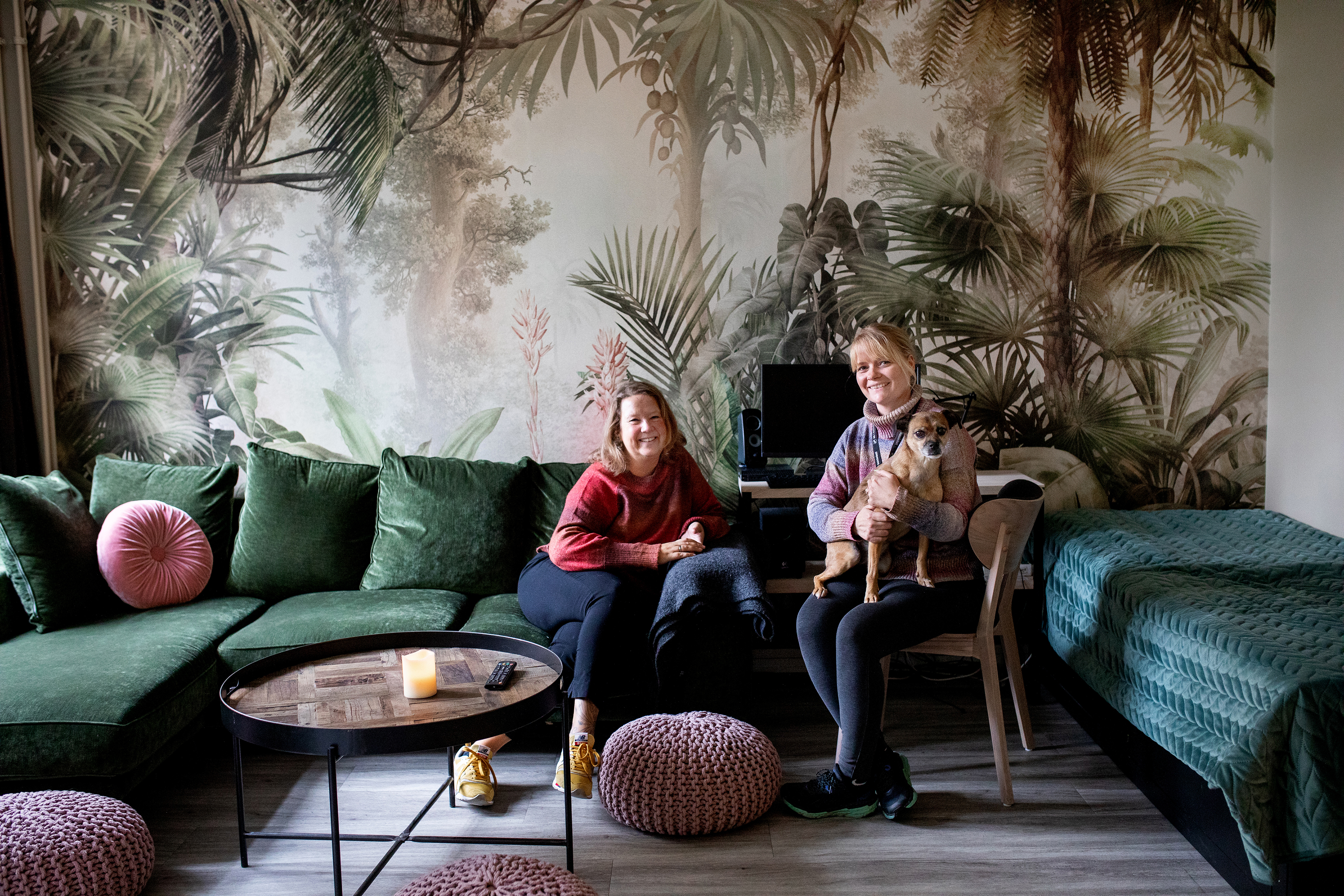 I en grønn velursofa foran en vegg med palmetapet sitter sosialfaglig leder Julie Haugsvold med hunden sin på fanget og ledende miljøterapaut Liv Solberg på Bosatt