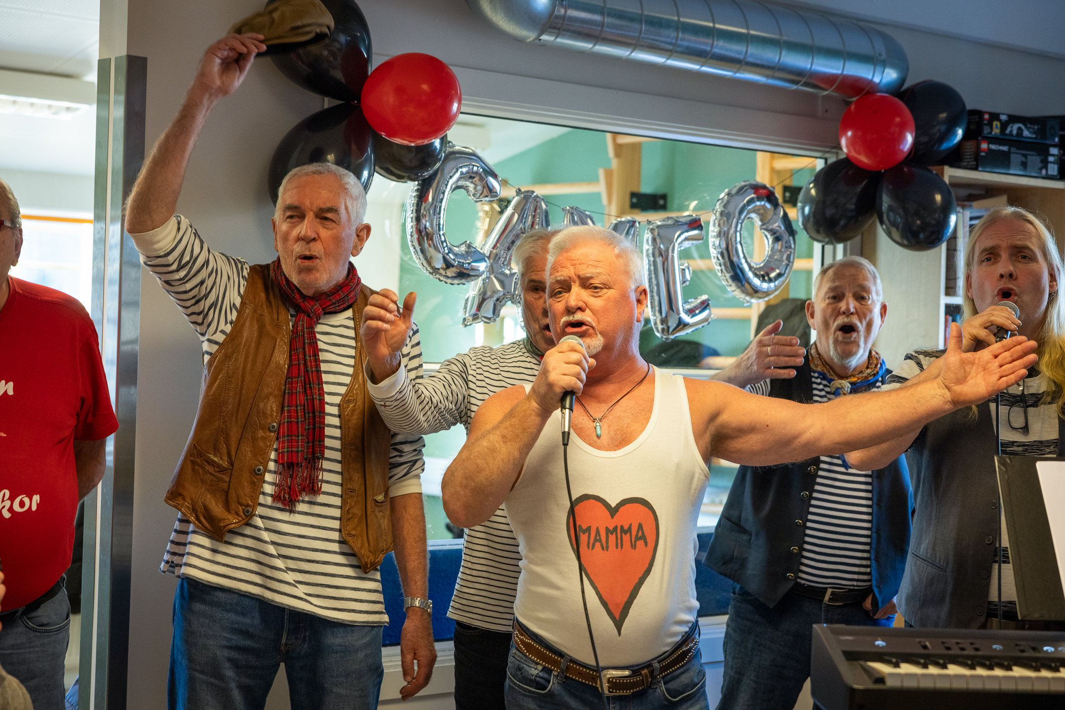 Fem seks voksne karer med stripete gensere og skjerf i halsen synger for full hals. En mann med singlet som det står Mamma på, holder en mikrofon. Bak er det pyntet med ballonger og oppblåsbare bokstaver som danner ordet CAMEO.