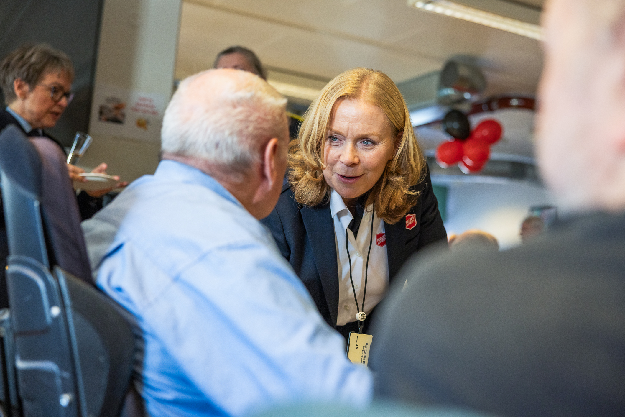 Middelaldrene kvinne i frelsesarmeuniform og halvlangt, lyst, rødlig hår snakker med en hvithåret eldre mann i lyseblå skjorte